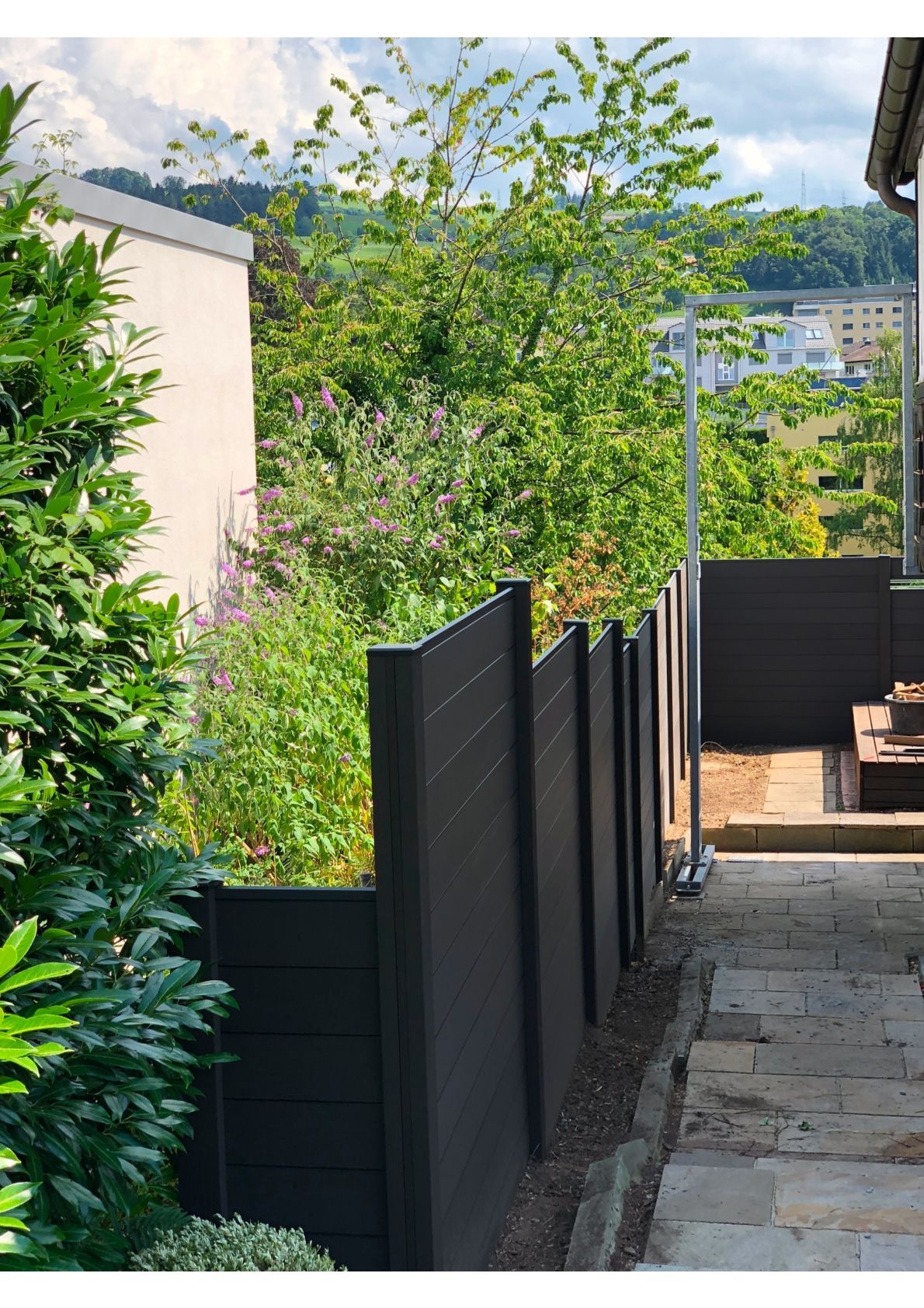 Schwarzer Zaun neben einem Gehweg, mit Grün und einem Gebäude im Hintergrund.