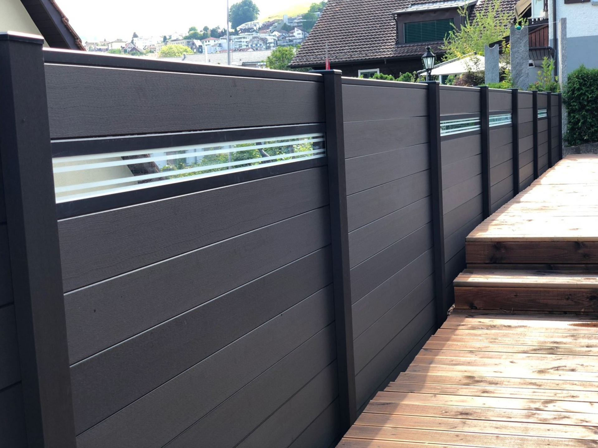 Dunkelgrauer moderner Zaun mit Glasplatten, Holzdeck und grünem Hintergrund.