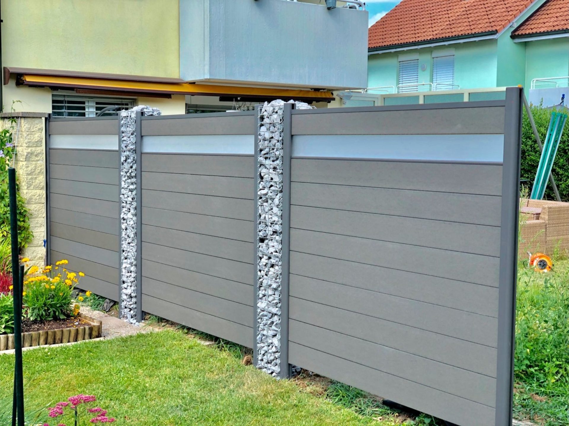 Grauer und steinerner Zaun in einem Hof mit horizontalen grauen Latten, Steinsäulen und einem hellgrauen horizontalen Band.