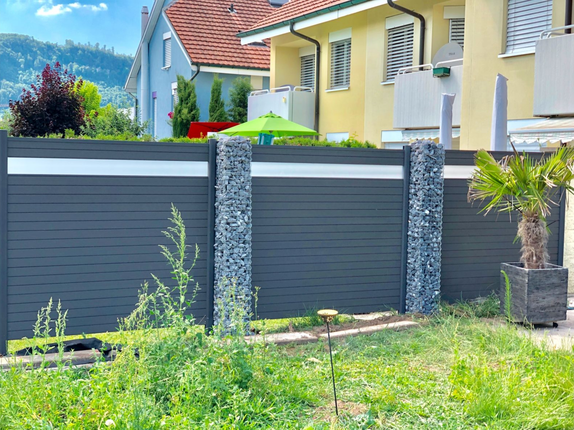 Dunkelgrauer Zaun mit Steinsäulen, Häuser im Hintergrund und eine kleine Palme auf der rechten Seite.