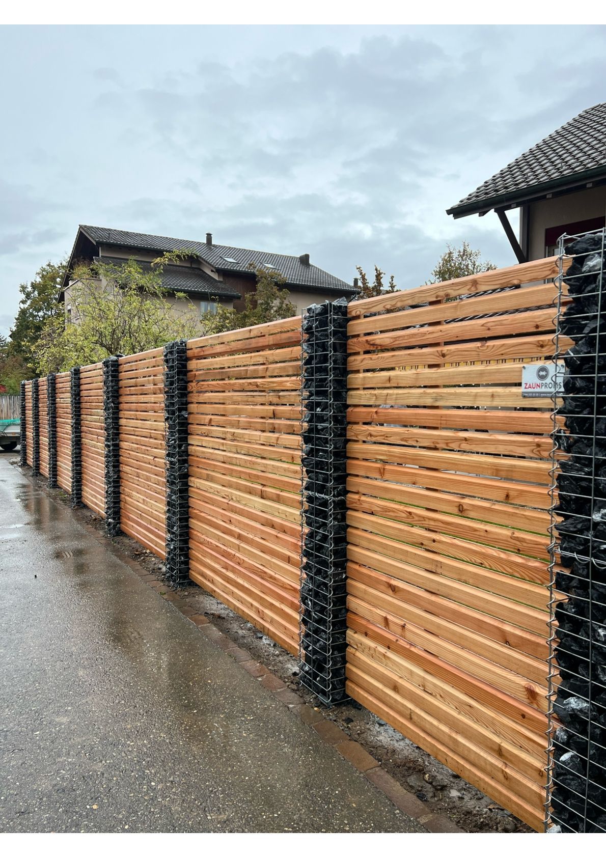 Holzzaun mit Steinsäulen, der an einer Straße entlangführt, bewölkter Tag.