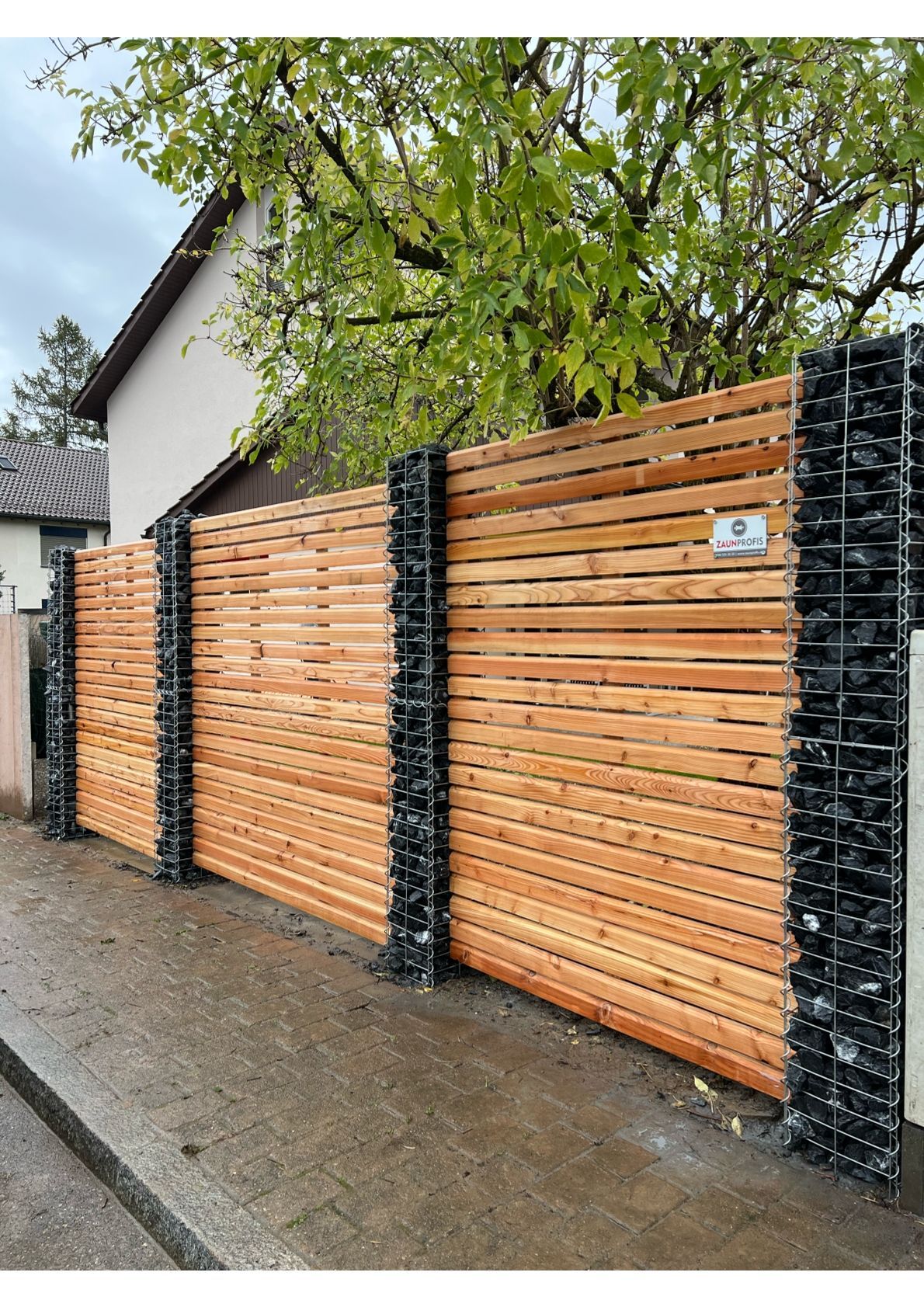 Holzzaun mit horizontalen Latten, flankiert von schwarzen Steinsäulen, entlang eines Gehwegs.