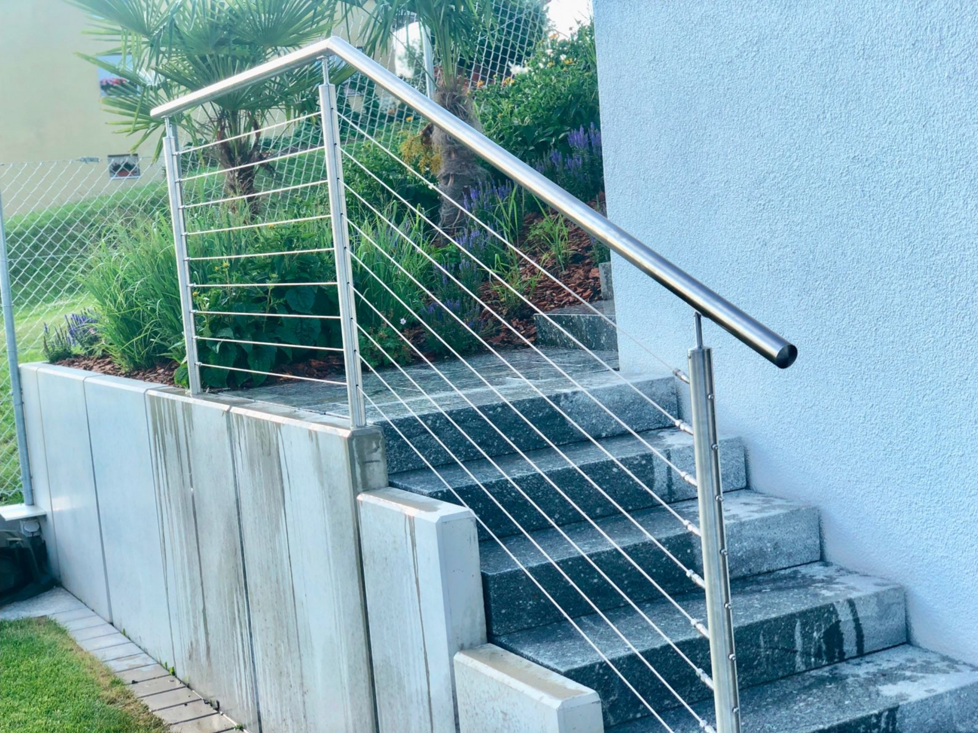 Edelstahlseilgeländer an Außentreppe und Stützmauer, im Hintergrund Pflanzen.