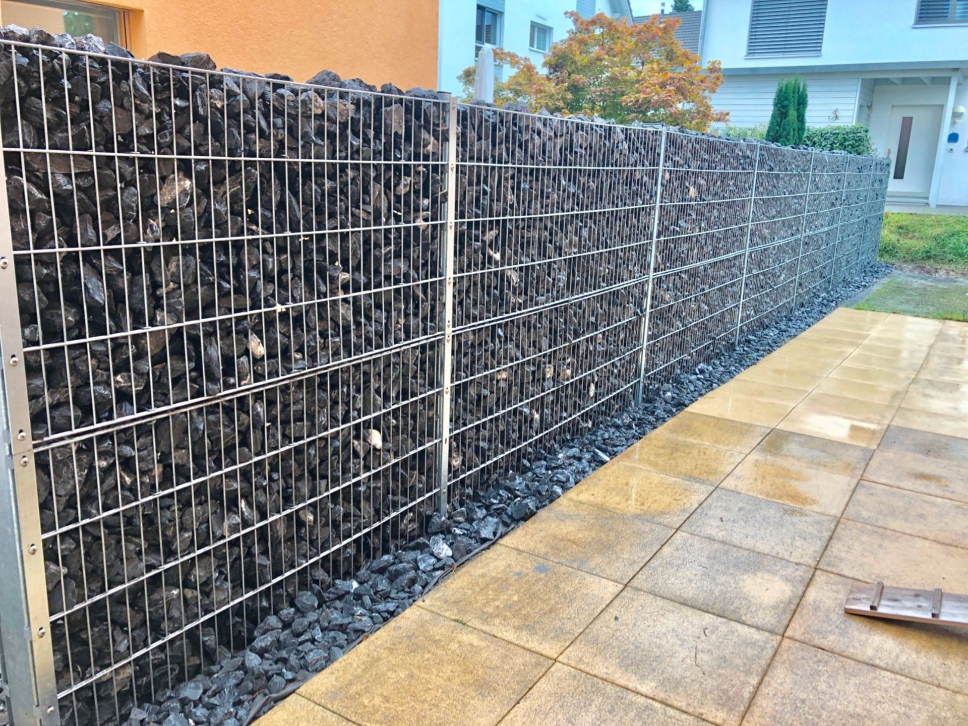 Mit dunklen Steinen gefüllte Gabionenwand, die an eine Terrasse mit hellen Pflastersteinen grenzt.