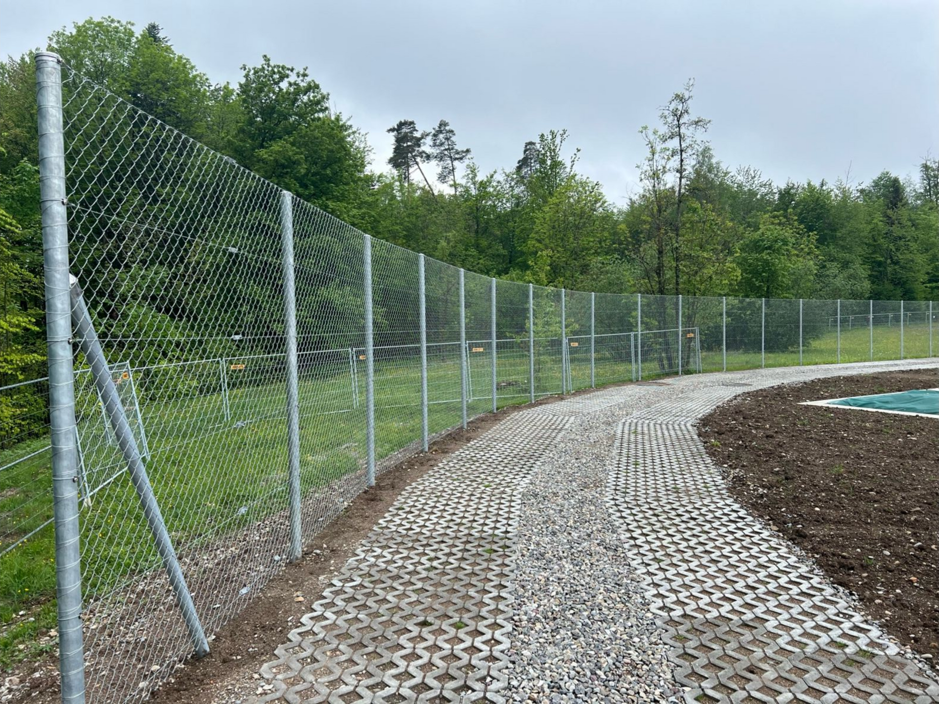Ein Maschendrahtzaun aus Metall umgibt einen Schotterweg durch ein Waldgebiet.