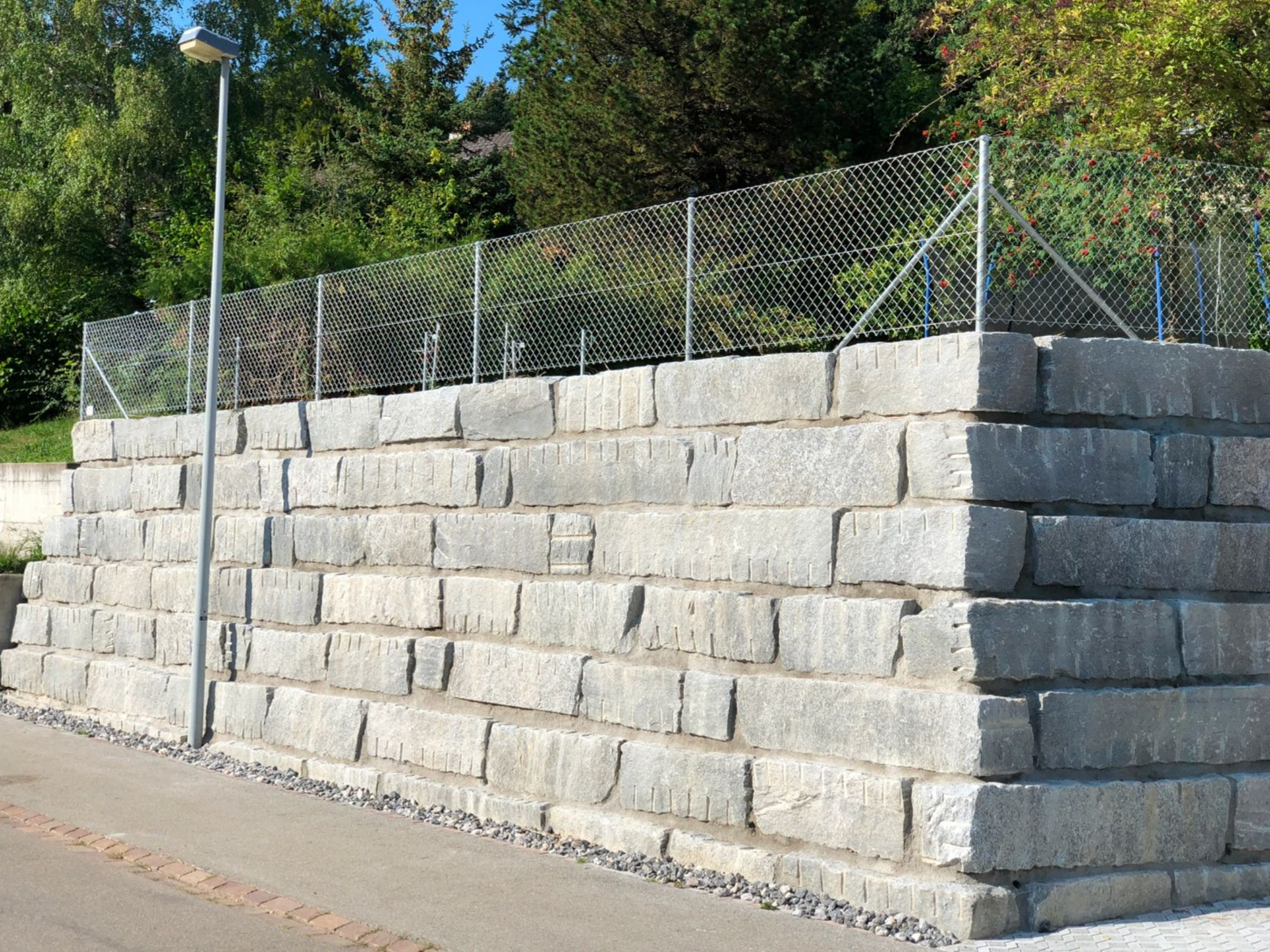 Steinstützmauer mit Maschendrahtzaun an einem leicht geneigten Hang mit einer Straße.