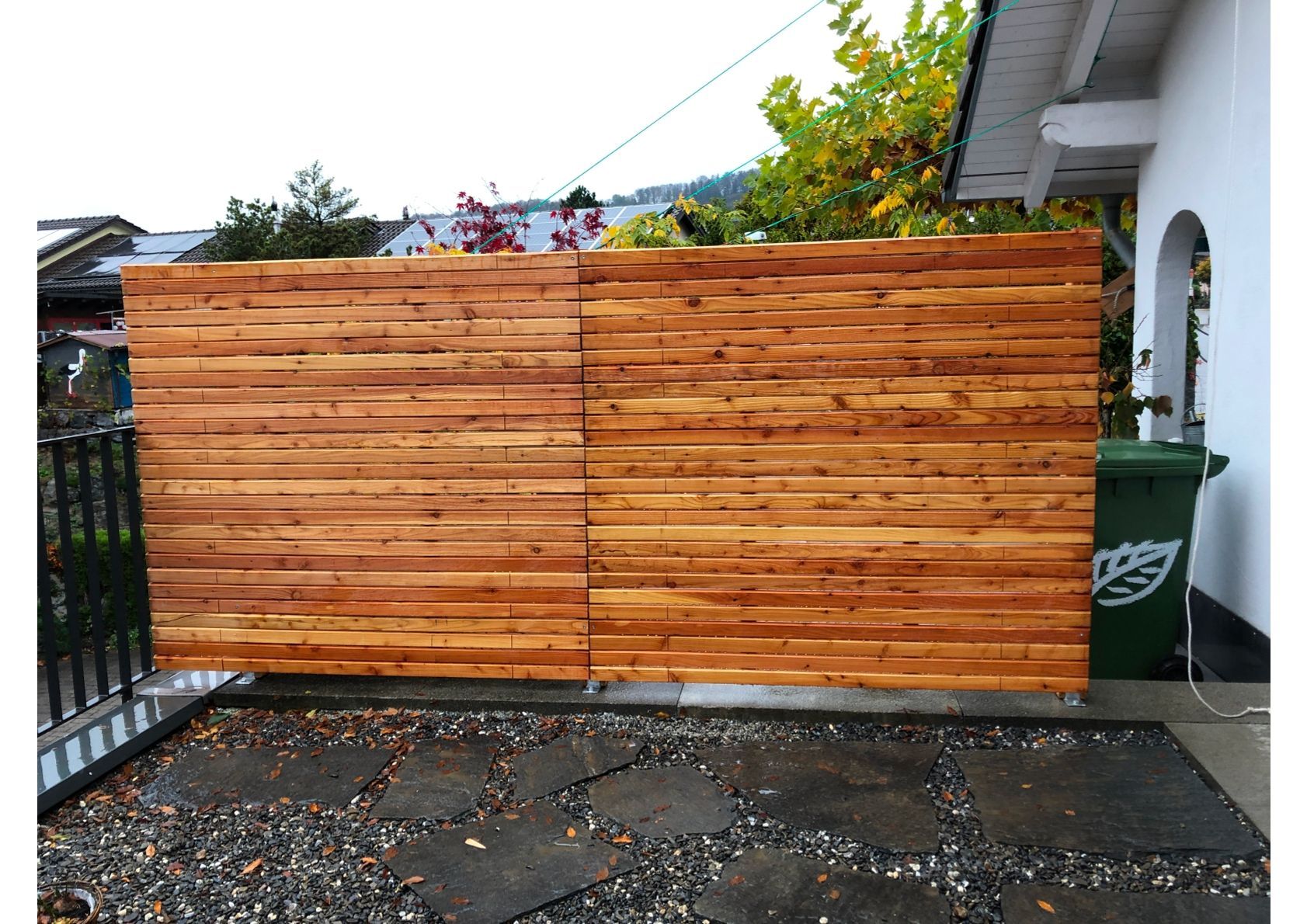 Sichtschutzzaun aus Holz auf einer Terrasse mit Steinpflaster, neben einem weißen Gebäude und einer grünen Mülltonne.