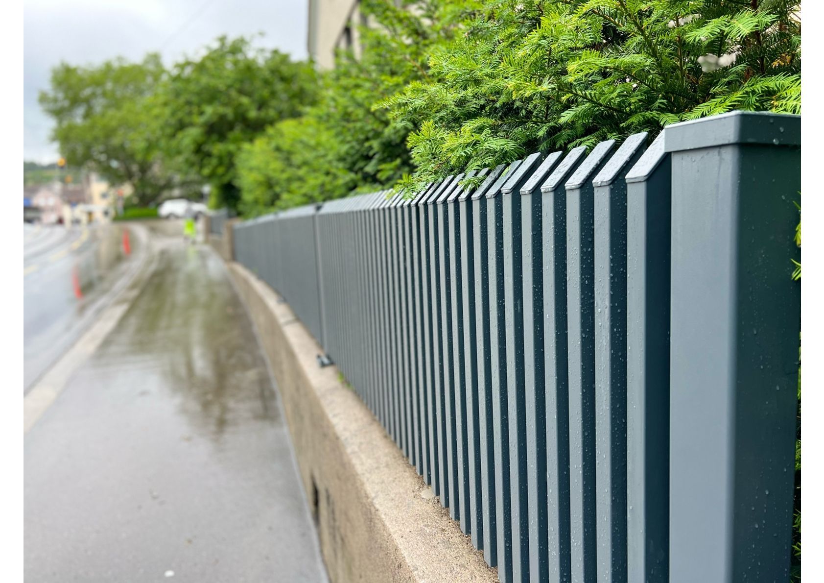 Grauer Metallzaun entlang eines nassen Gehwegs neben einer Straße, mit grünem Laub im Hintergrund.