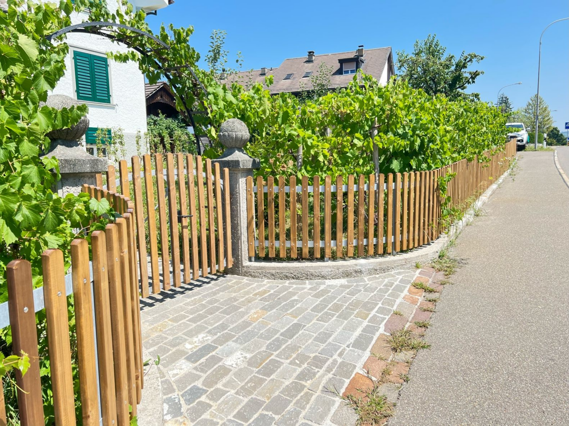 Holzzaun vor einem Haus mit Weinreben und Kopfsteinpflasterweg an einem sonnigen Tag.