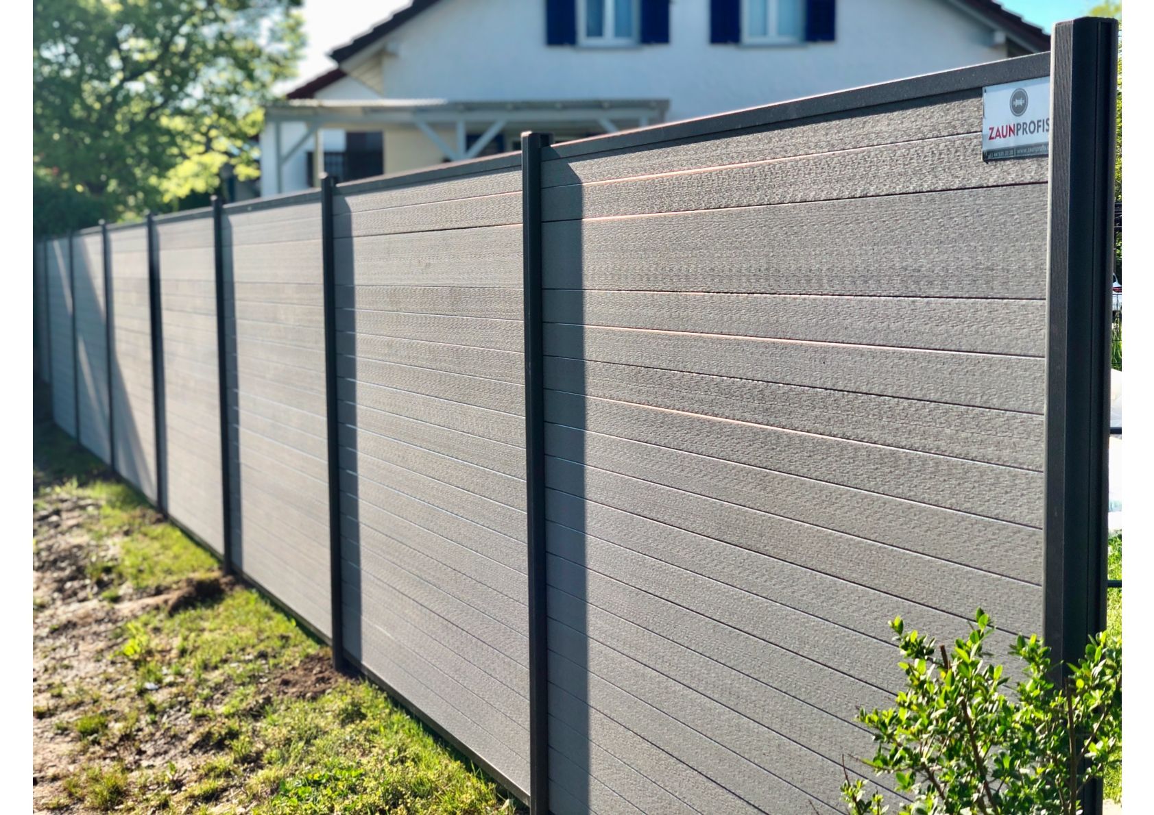 Grauer Sichtschutzzaun mit schwarzen Pfosten vor einem Haus.