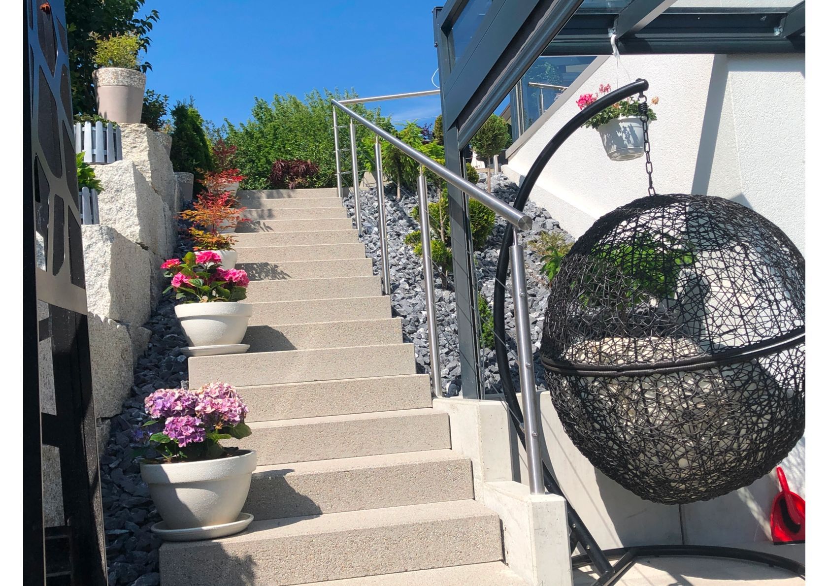 Steinstufen mit Blumen in weißen Töpfen, ein Metallgeländer und ein Hängesessel im Freien.