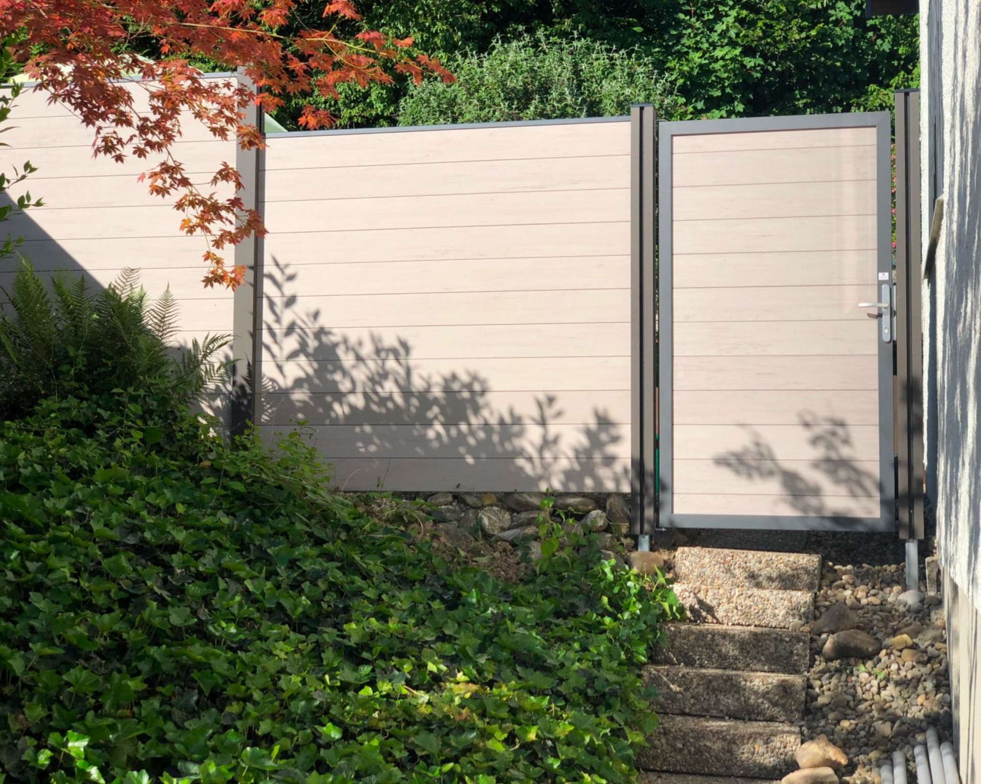 Beiger Zaun mit Tor in einer Gartenanlage, neben einer Steintreppe, umgeben von Grün.
