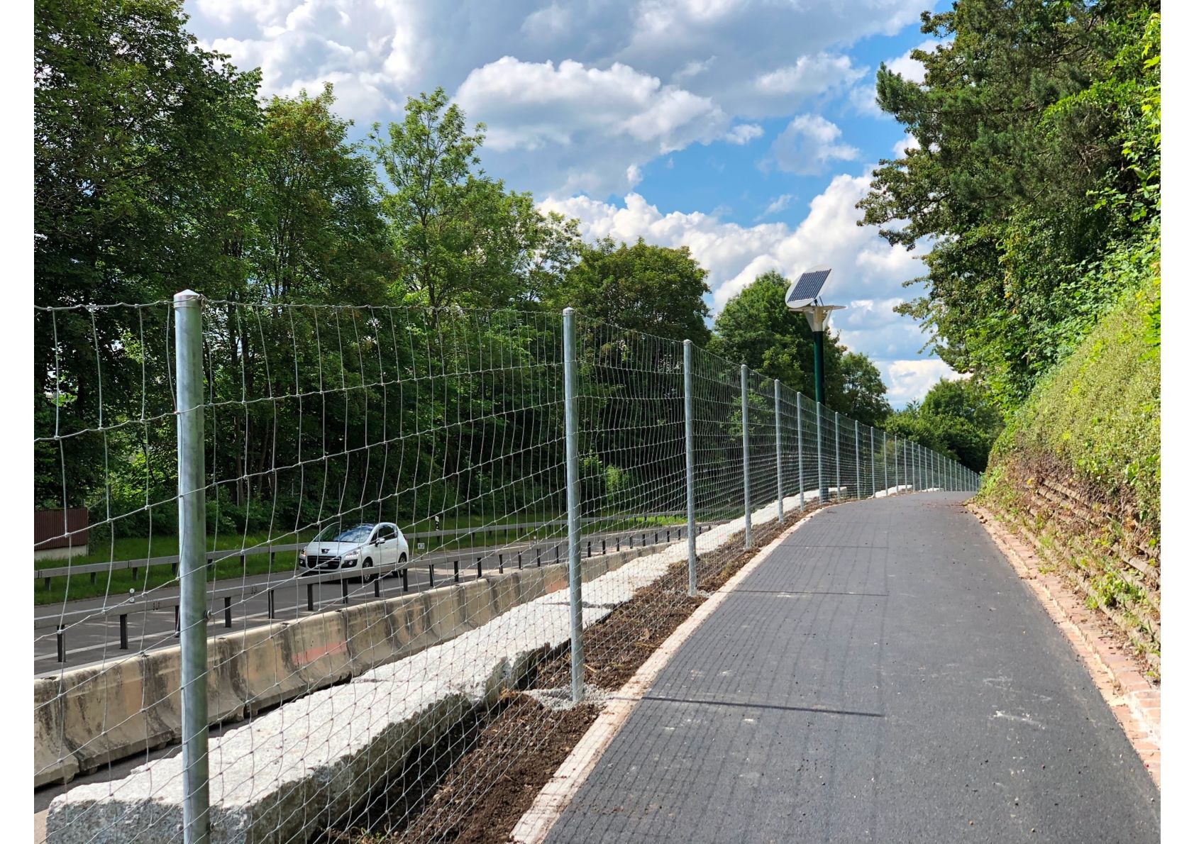 Ein gepflasterter Weg verläuft neben einer Straße und einem Zaun. Autos und Bäume sind sichtbar. Bewölkter Himmel.