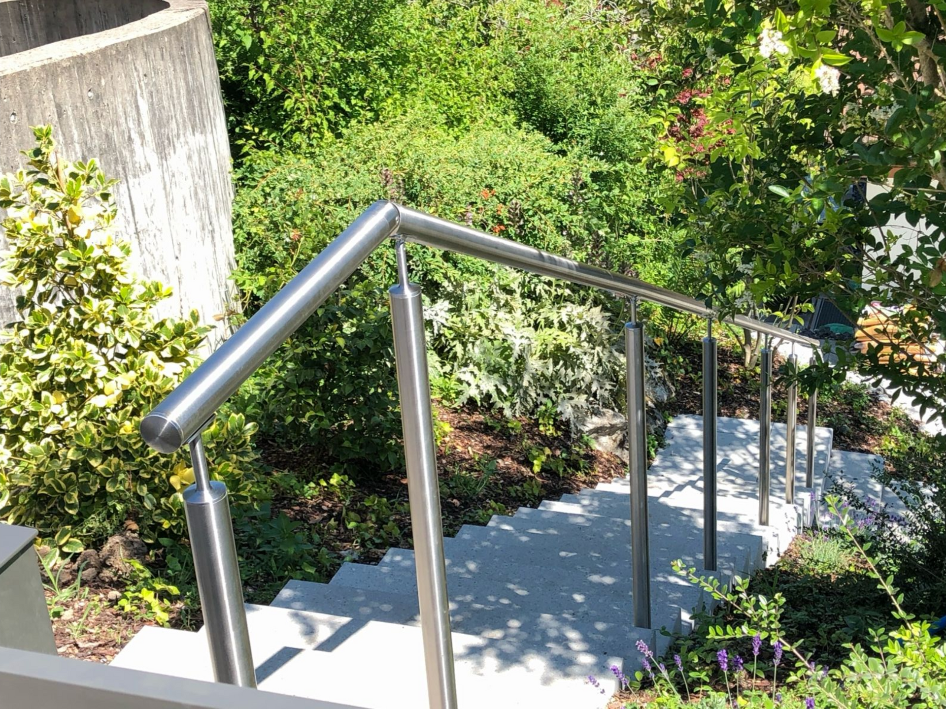 Treppe mit Edelstahlgeländer, die hinunter in einen üppigen Garten mit grünem Laub führt.