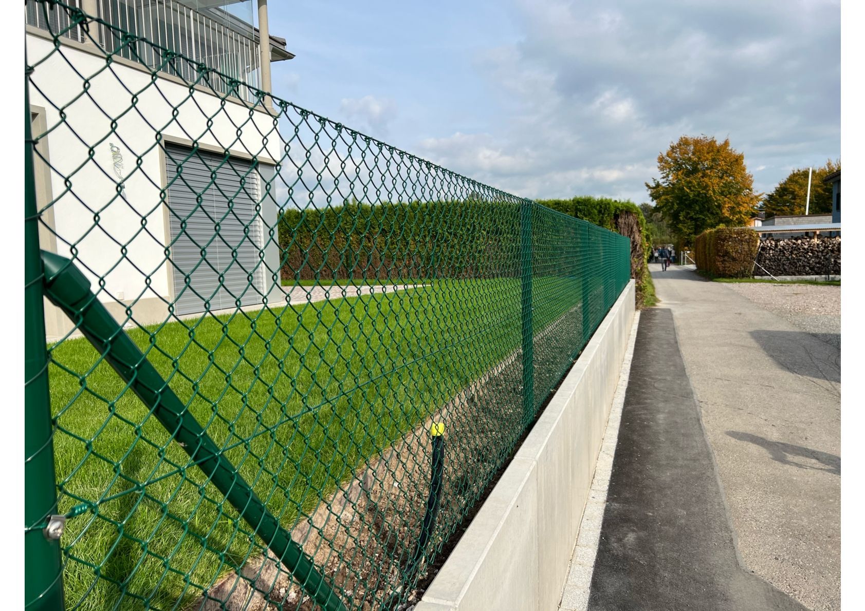 Grüner Maschendrahtzaun mit einer Hecke dahinter, neben einem Gehweg.