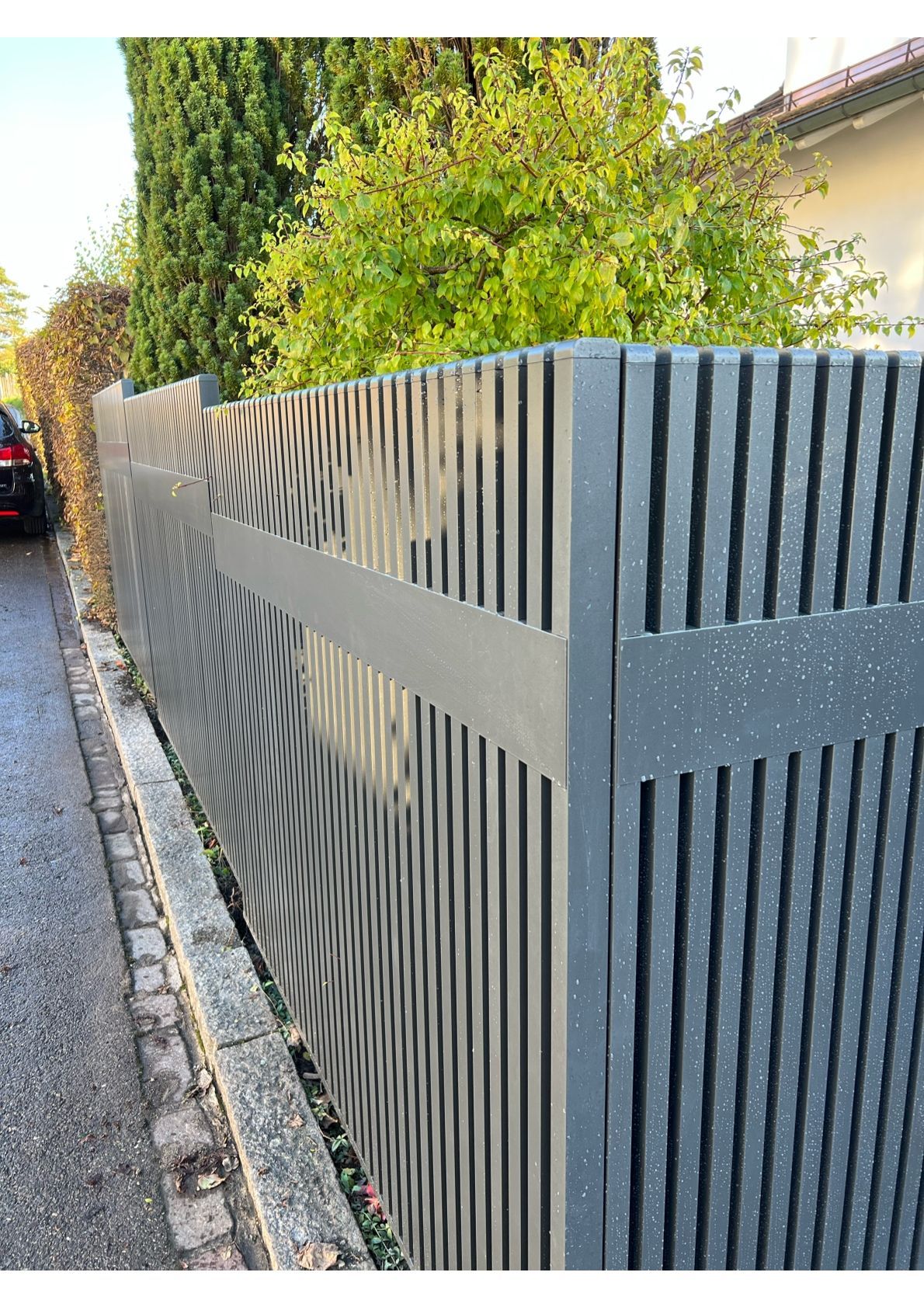 Grauer vertikaler Lattenzaun entlang eines Gehwegs mit Bäumen und einem Haus im Hintergrund.