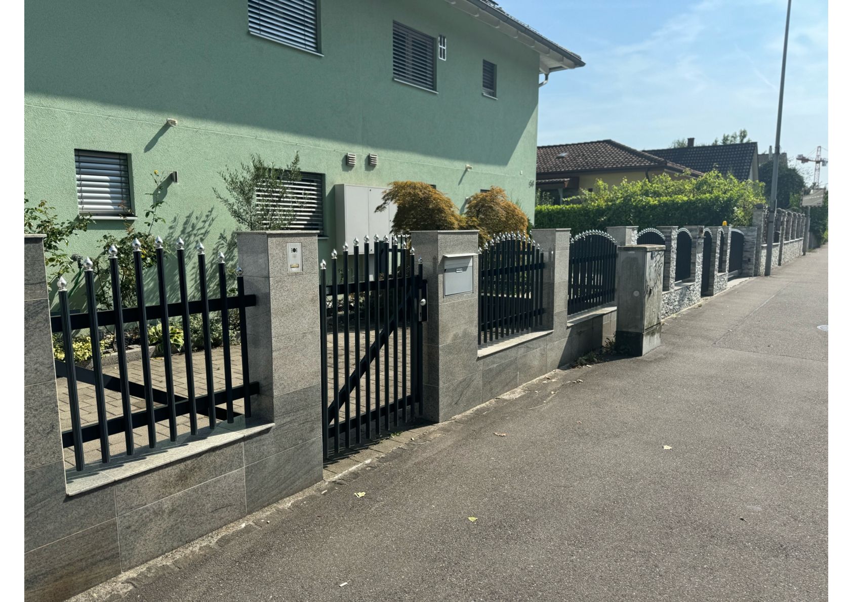 Grünes Haus mit schwarzem Eisenzaun und Tor, auf einer grauen Betonwand.