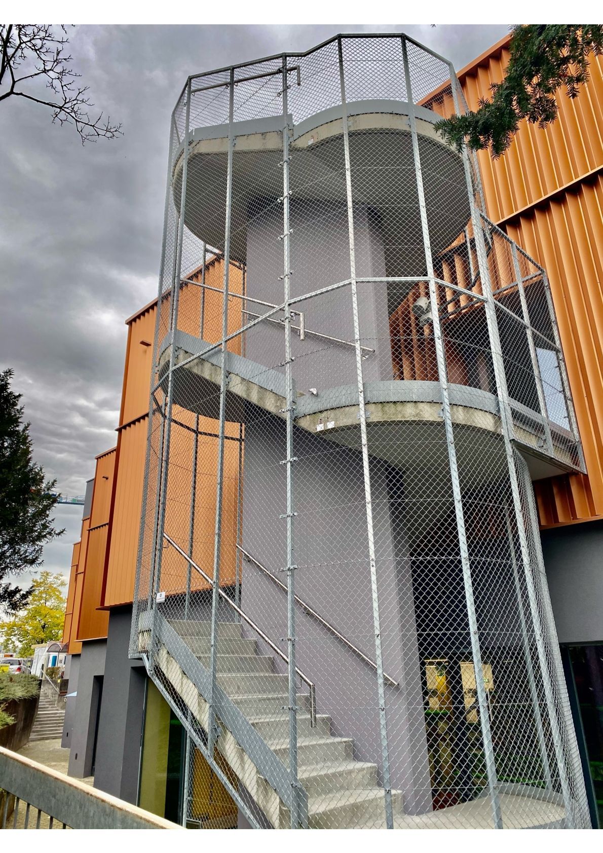 Aussenansicht einer Metalltreppe mit Netz, die an einem Gebäude mit orangefarbener Außenverkleidung angebracht ist.