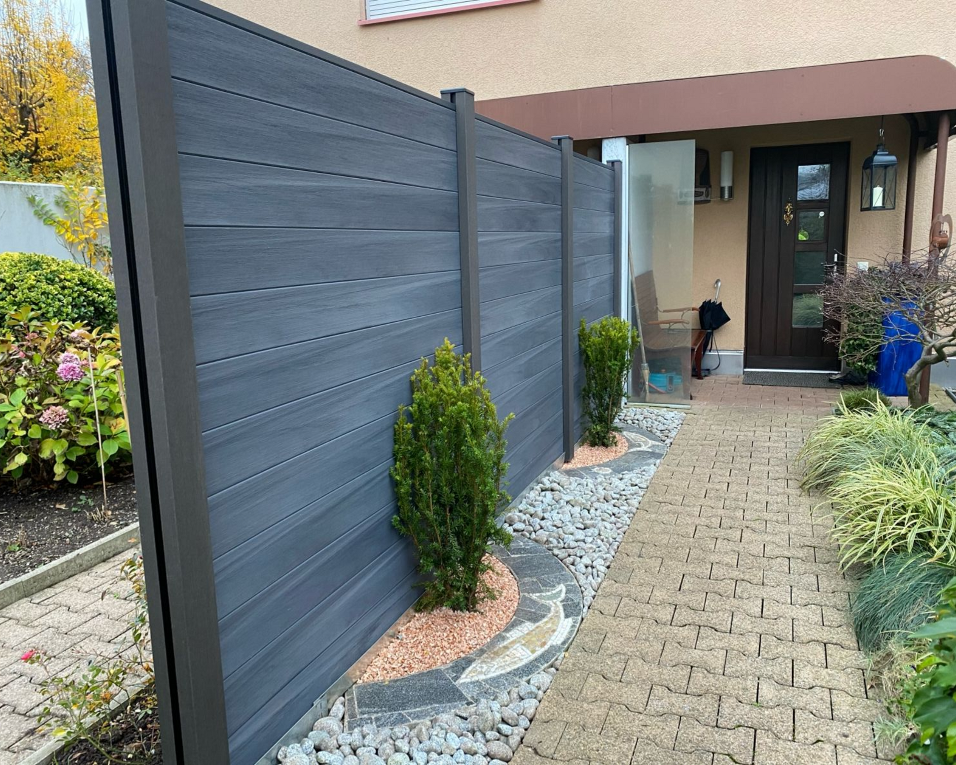 Dunkelgrauer Sichtschutzzaun entlang eines gemauerten Gehwegs mit grünen Büschen und Landschaftsgestaltung.