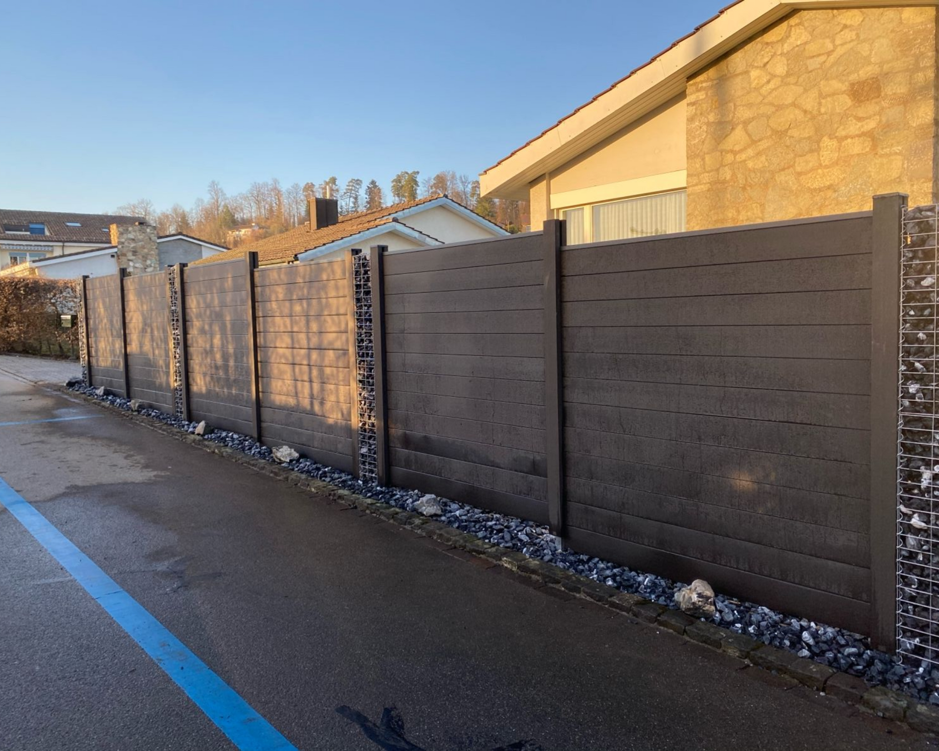 Dunkler Holzzaun entlang einer Straße, neben einem Haus mit Steinfassade.