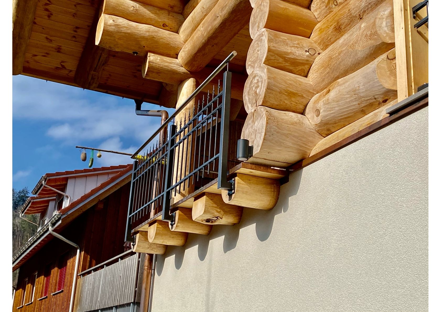 Blockhausbalkon mit Metallgeländer vor heller Stuckwand und klarem Himmel.