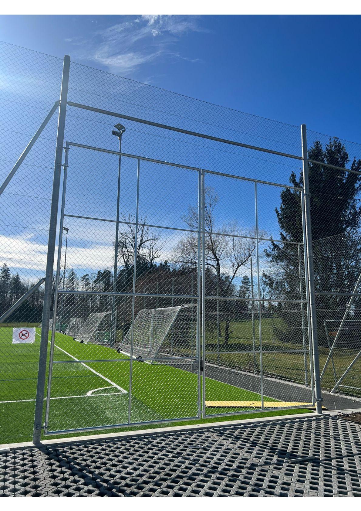 Metalltor und Zaun umgeben ein Fußballfeld mit grünem Gras und Toren. Klarer blauer Himmel.