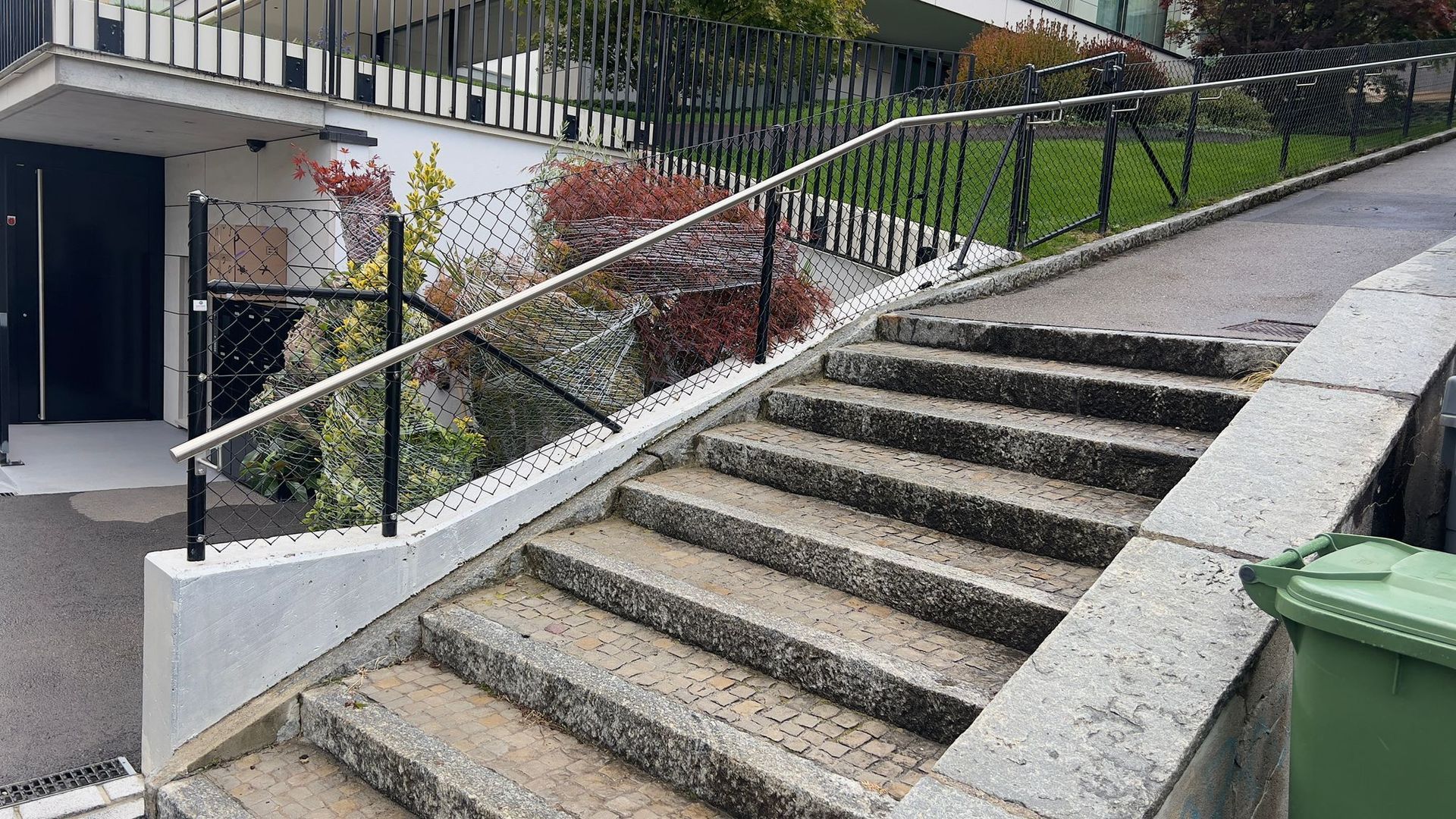 Steintreppe, die bergauf führt, mit schwarzem Geländer und einem grünen Mülleimer auf der rechten Seite.