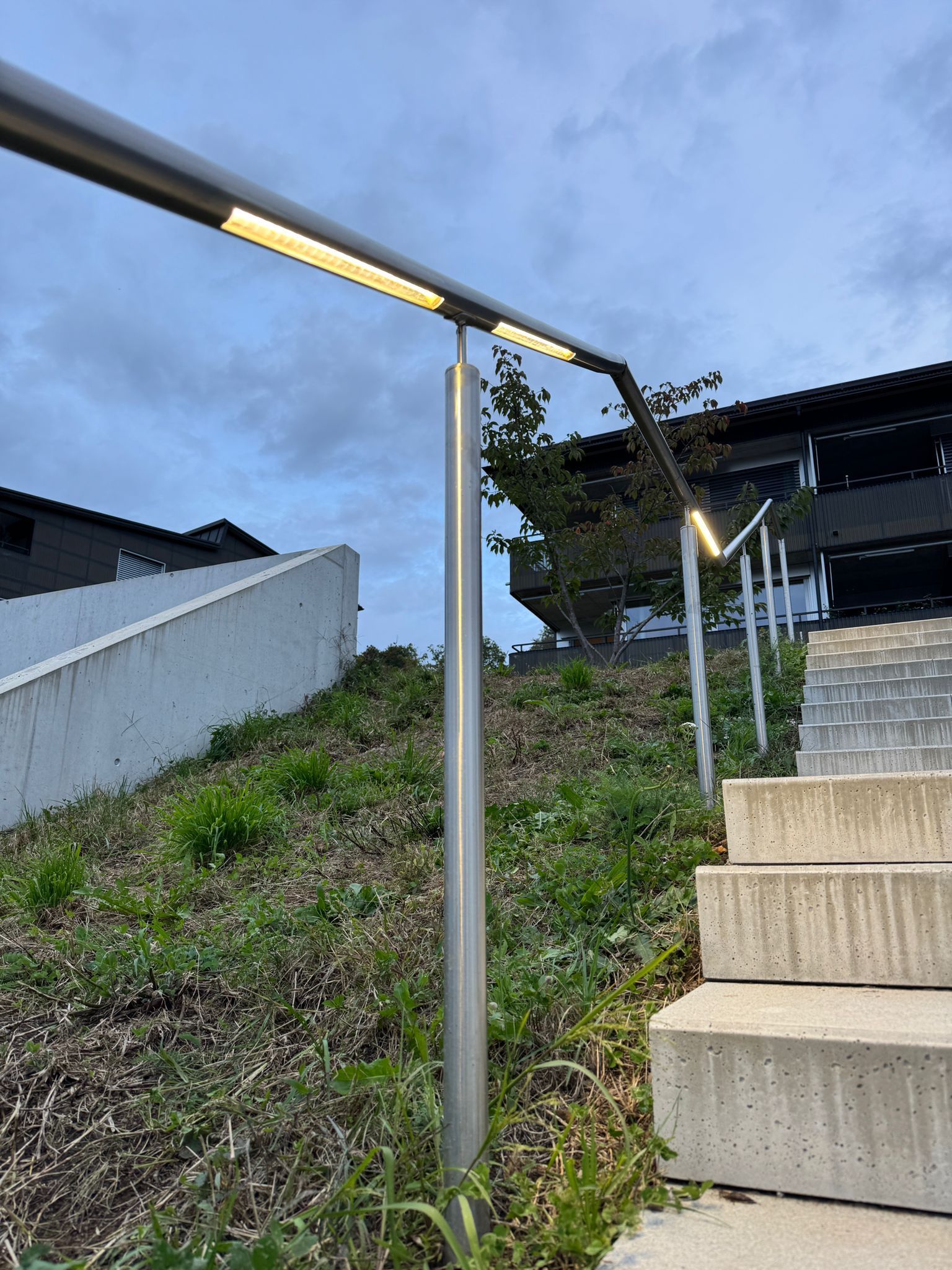 Treppe mit beleuchtetem Metallhandlauf. Betonstufen führen einen grasbewachsenen Hang hinauf; im Hintergrund Gebäude.