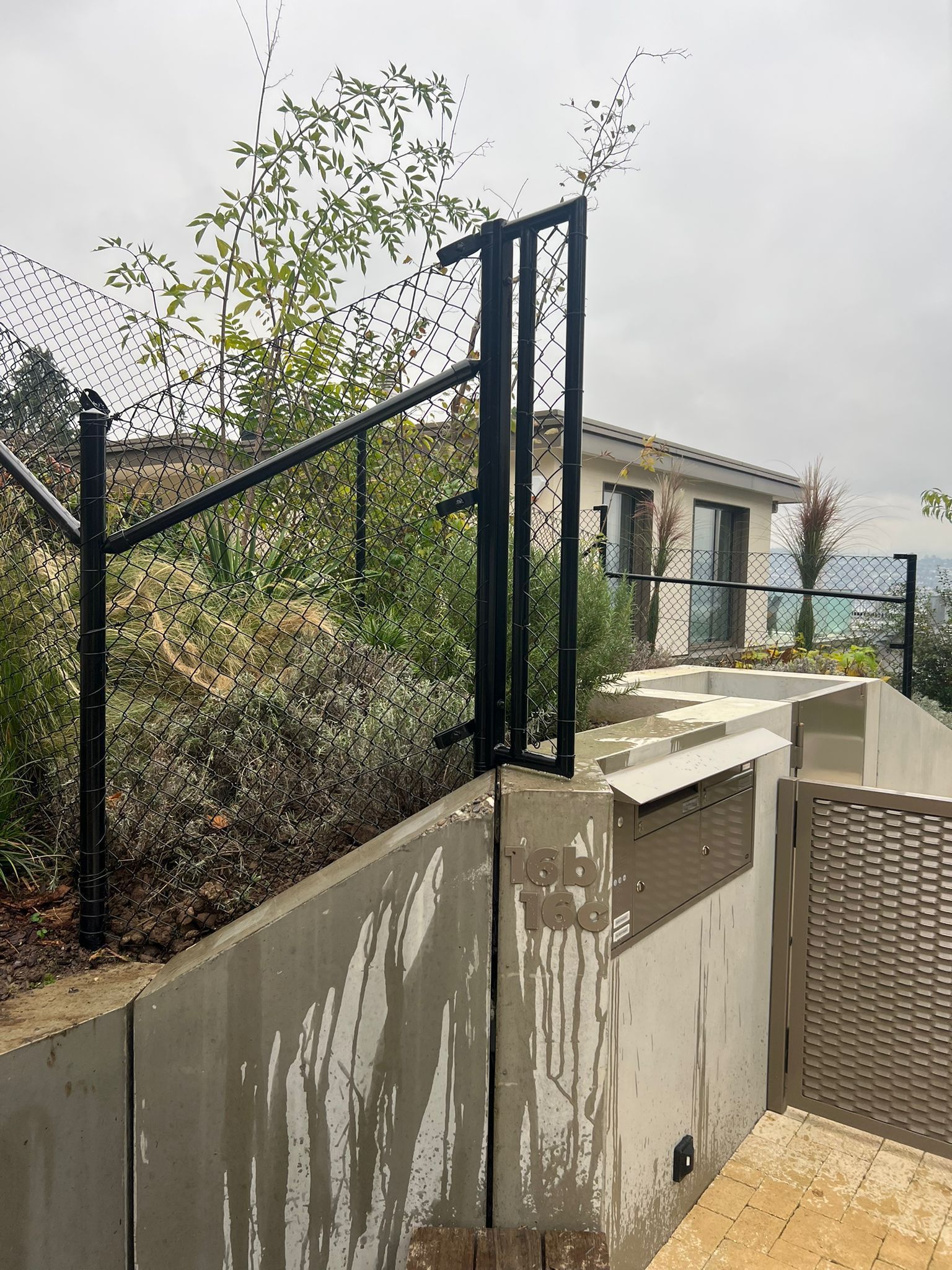 Schwarzes Metallgeländer an einer Betonwand, Grünflächen und im Hintergrund ein sichtbares Haus.