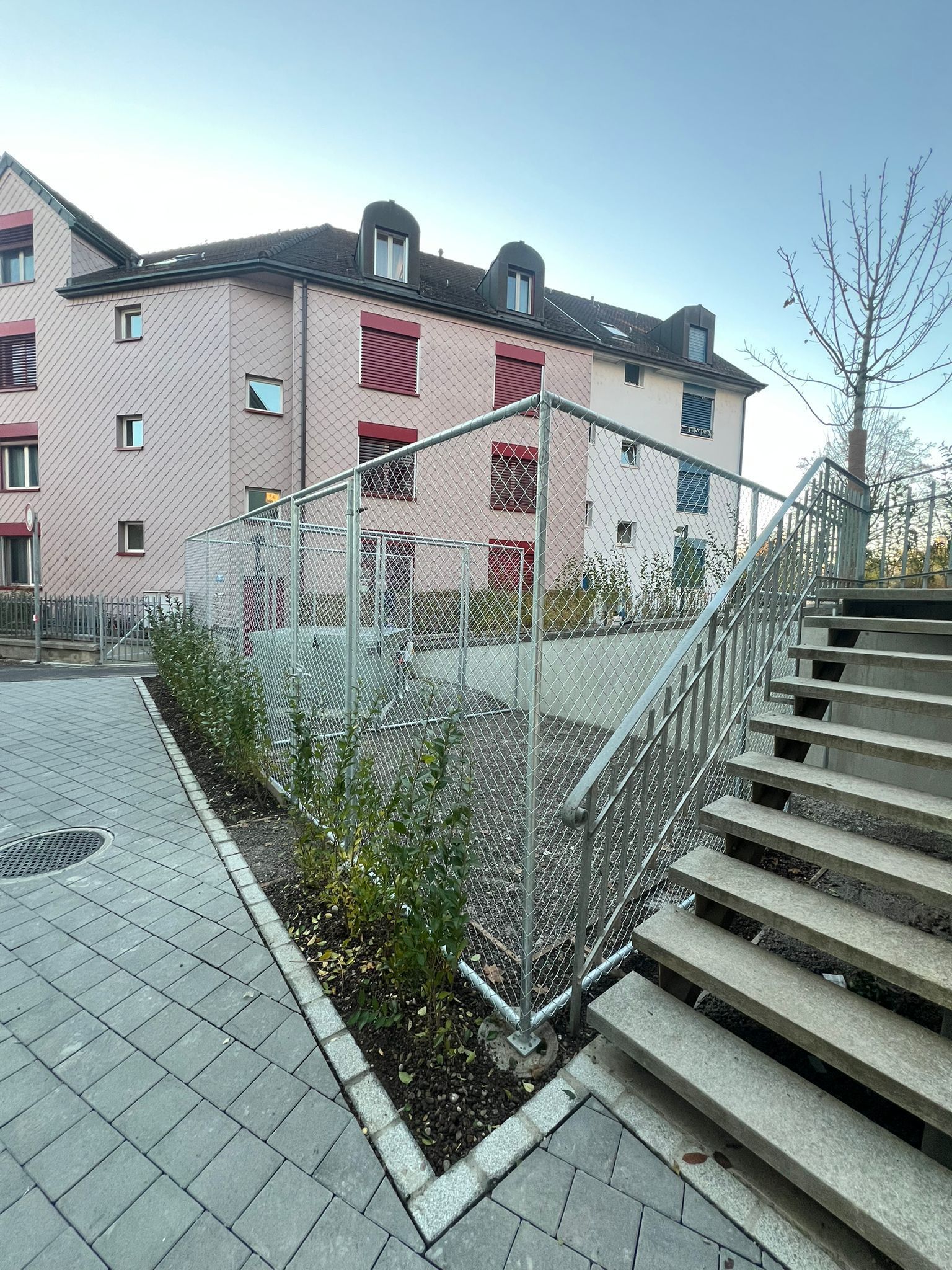 Rosafarbenes Gebäude mit roten Fensterläden hinter einem Metallzaun, Treppen und einem gepflasterten Gehweg.