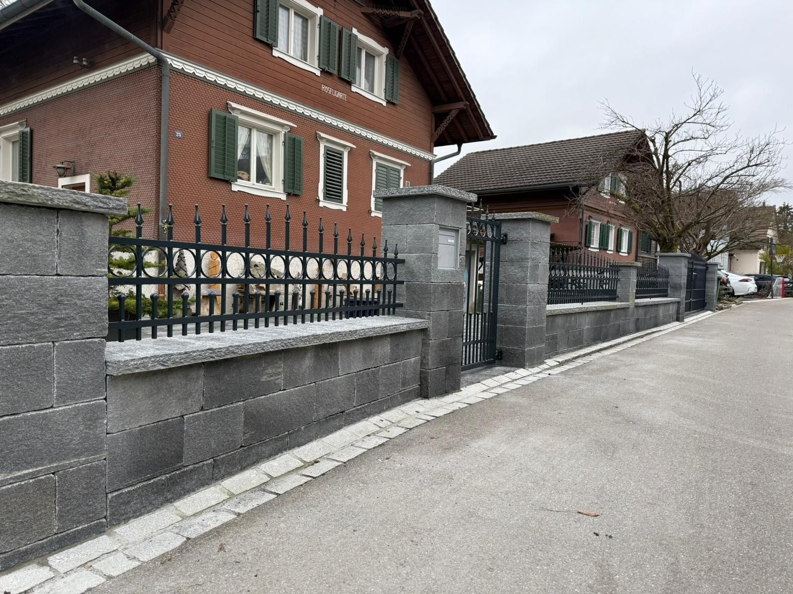 Steinmauer mit schwarzem Metallzaun entlang einer Straße vor einem Backsteinhaus.