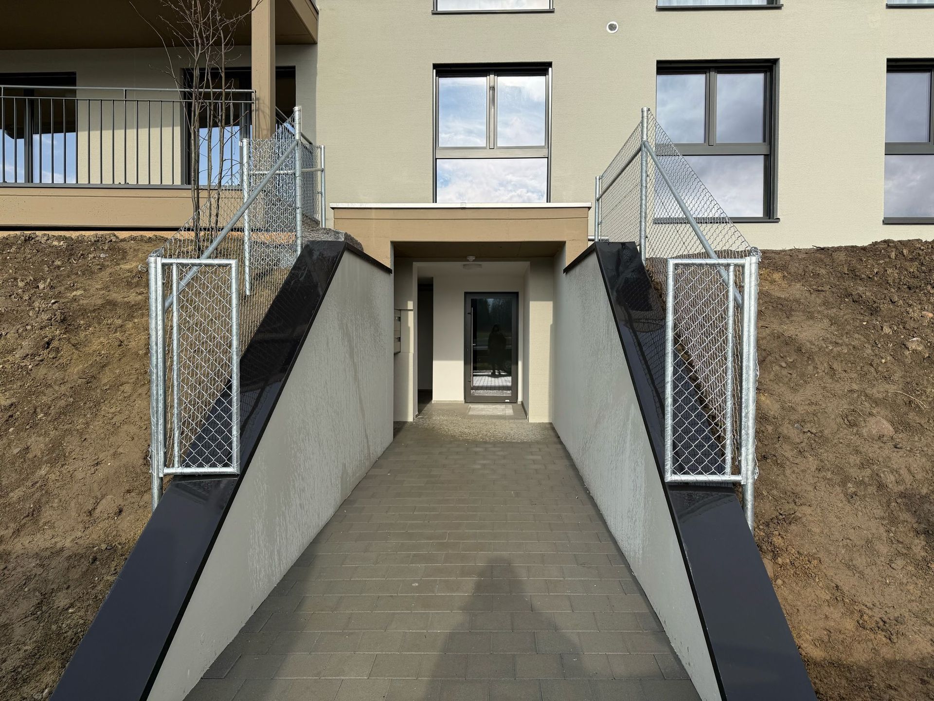 Eingang mit Betonrampe, die zu einer Tür führt, flankiert von Metallgeländern und Betonwänden.