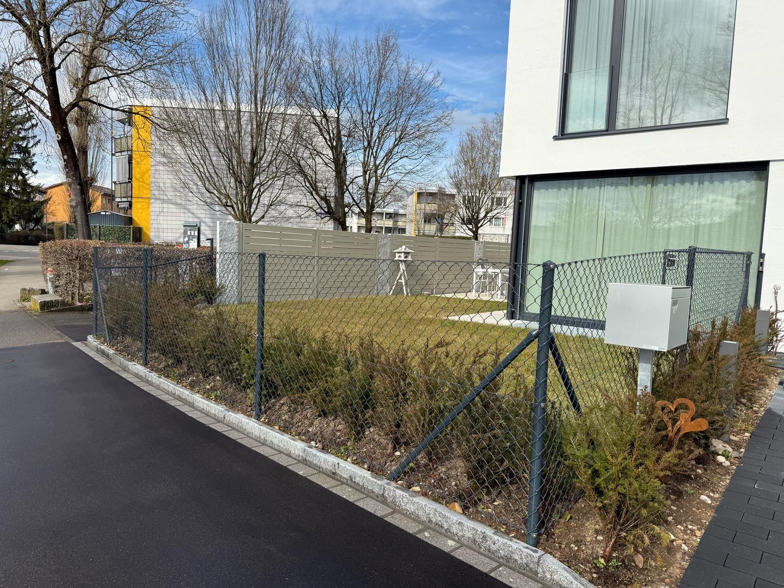 An einem sonnigen Tag umschließt ein grauer Zaun einen Hof mit Büschen und einem modernen weißen Gebäude im Hintergrund.