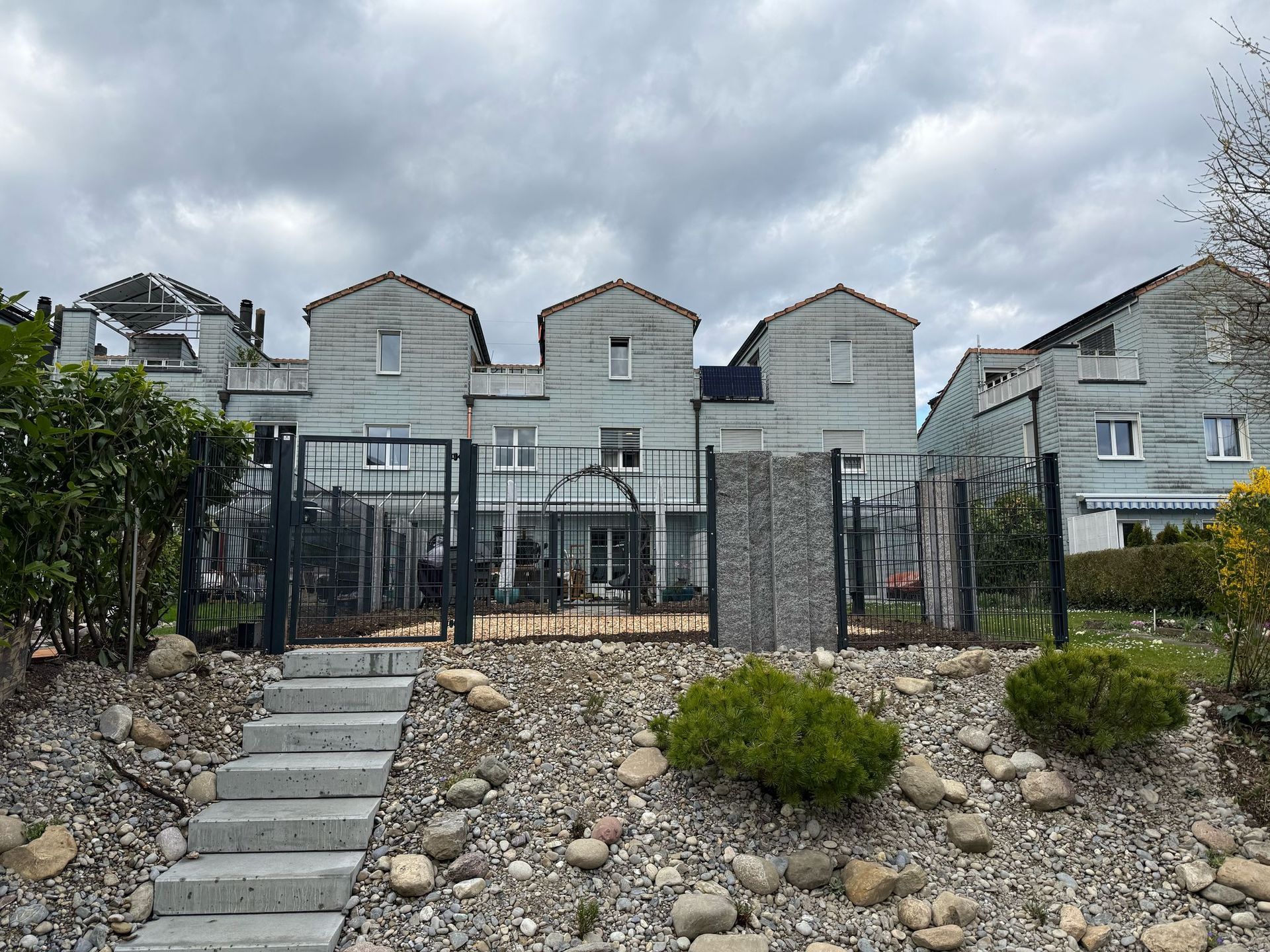 Reihe grauer Häuser mit Steinstufen, Zaun und bewölktem Himmel.