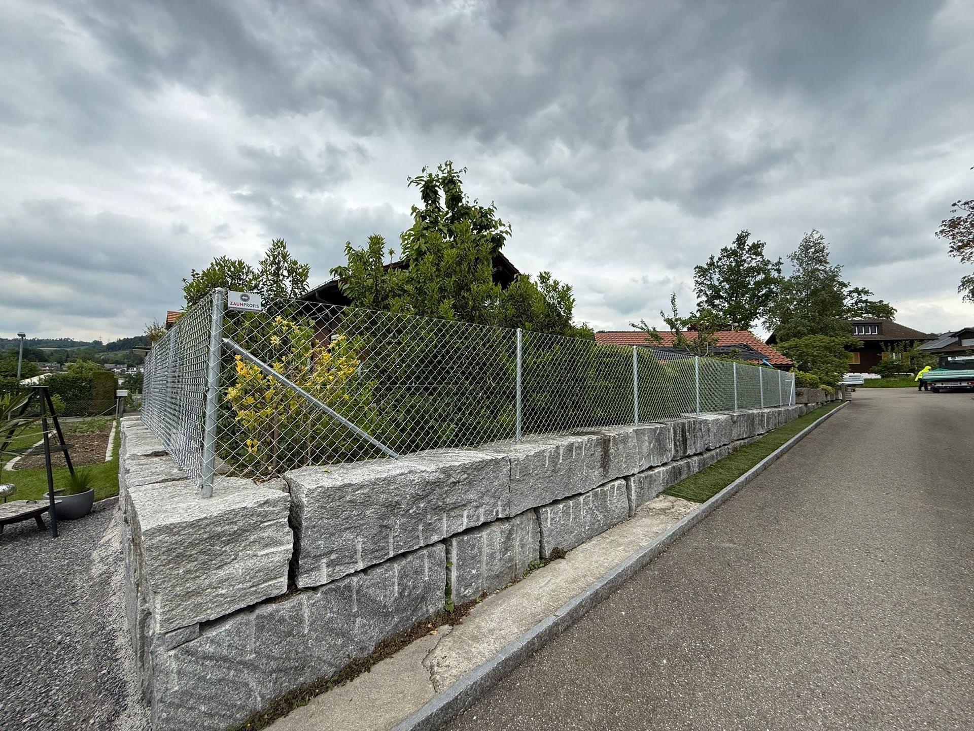 Steinmauer mit Maschendrahtzaun, ein Haus, Bäume und eine Straße unter einem bewölkten Himmel.