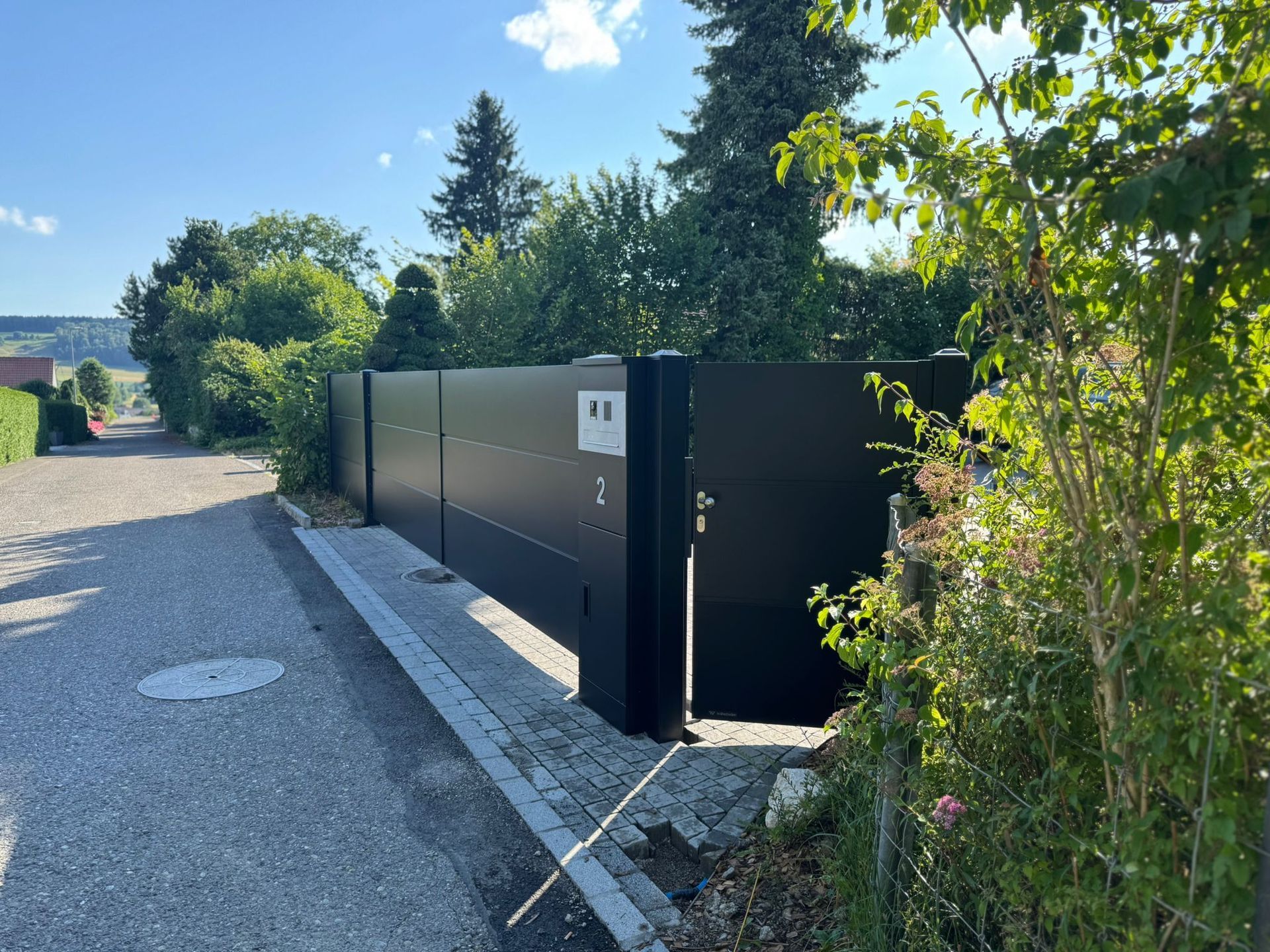 Schwarzes Metalltor auf einem Schotterweg, der zu einem Wohngebiet mit Bäumen unter blauem Himmel führt.