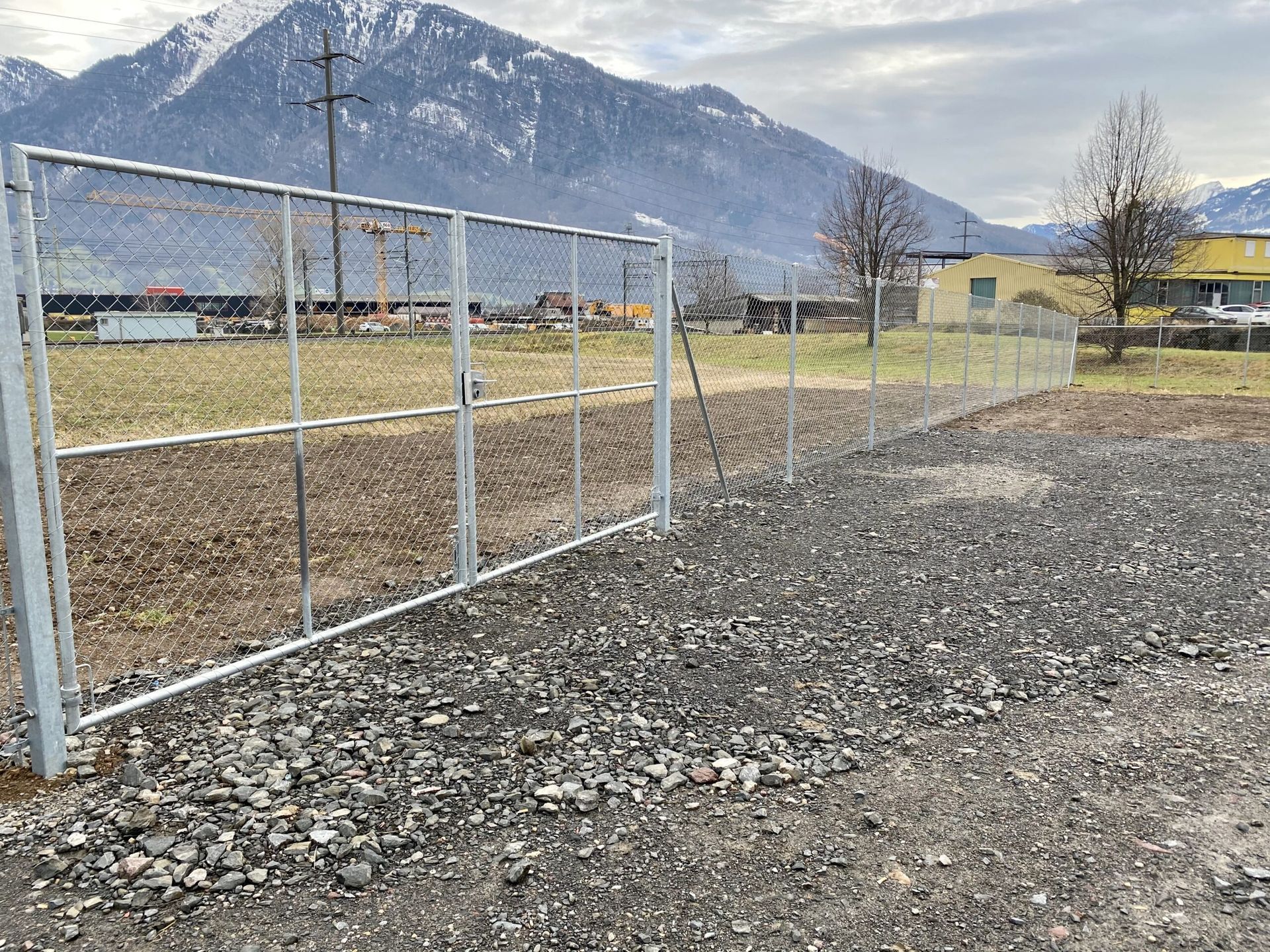 Metallischer Maschendrahtzaun mit offenem Tor in einem Kiesgebiet; Berge im Hintergrund.