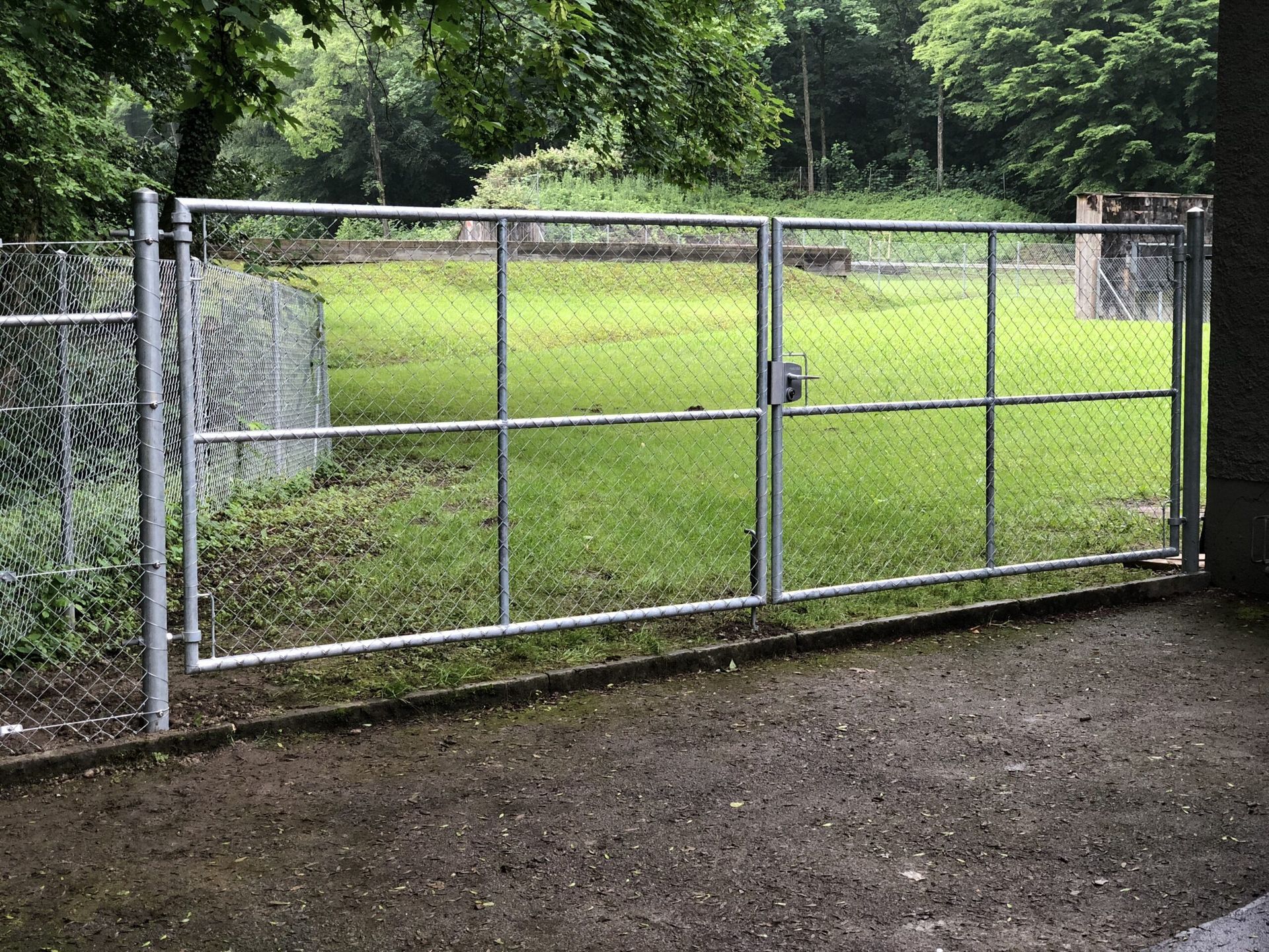 Ein Maschendrahtzaun mit einem Doppeltor auf einer Rasenfläche, im Hintergrund Bäume.