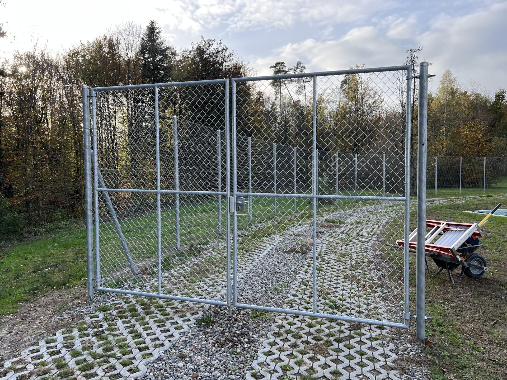 Metallisches Maschendrahttor vor einem Waldstück auf einer Kiesfläche.