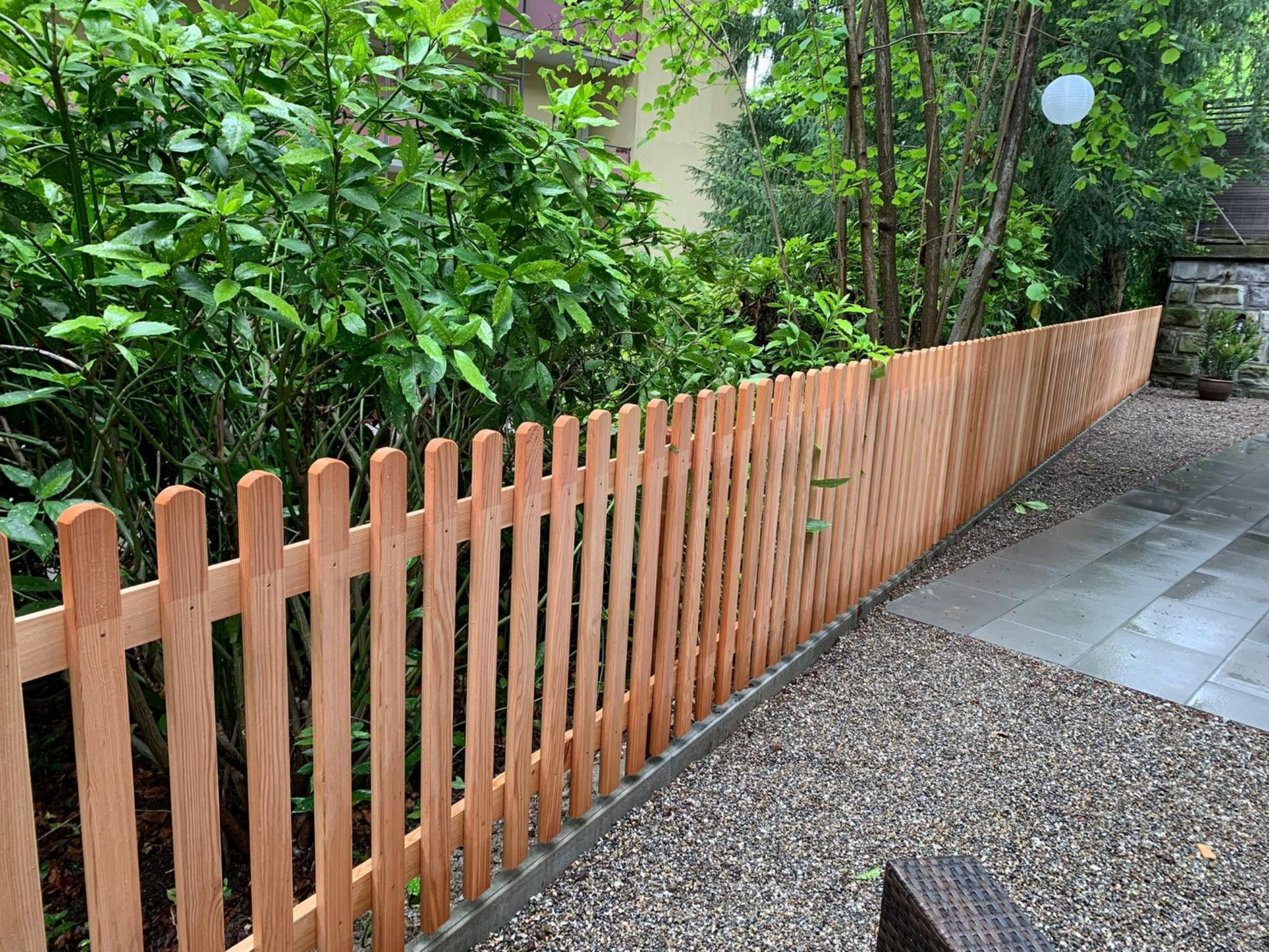 Holzlattenzaun entlang einer Steinterrasse und einer Kiesgrenze, mit grünen Büschen im Hintergrund.