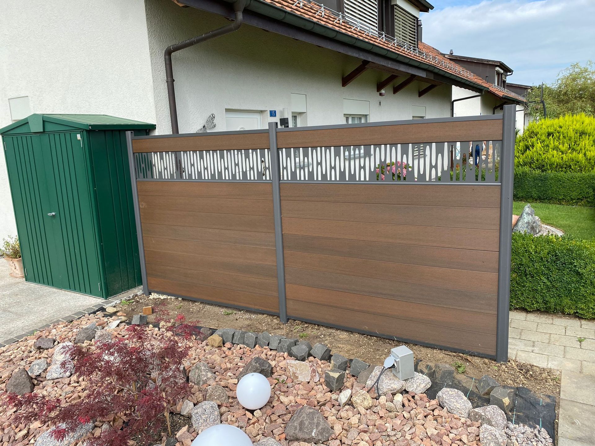 Braun-grauer Verbundzaun mit dekorativen oberen Paneelen vor einem Haus.