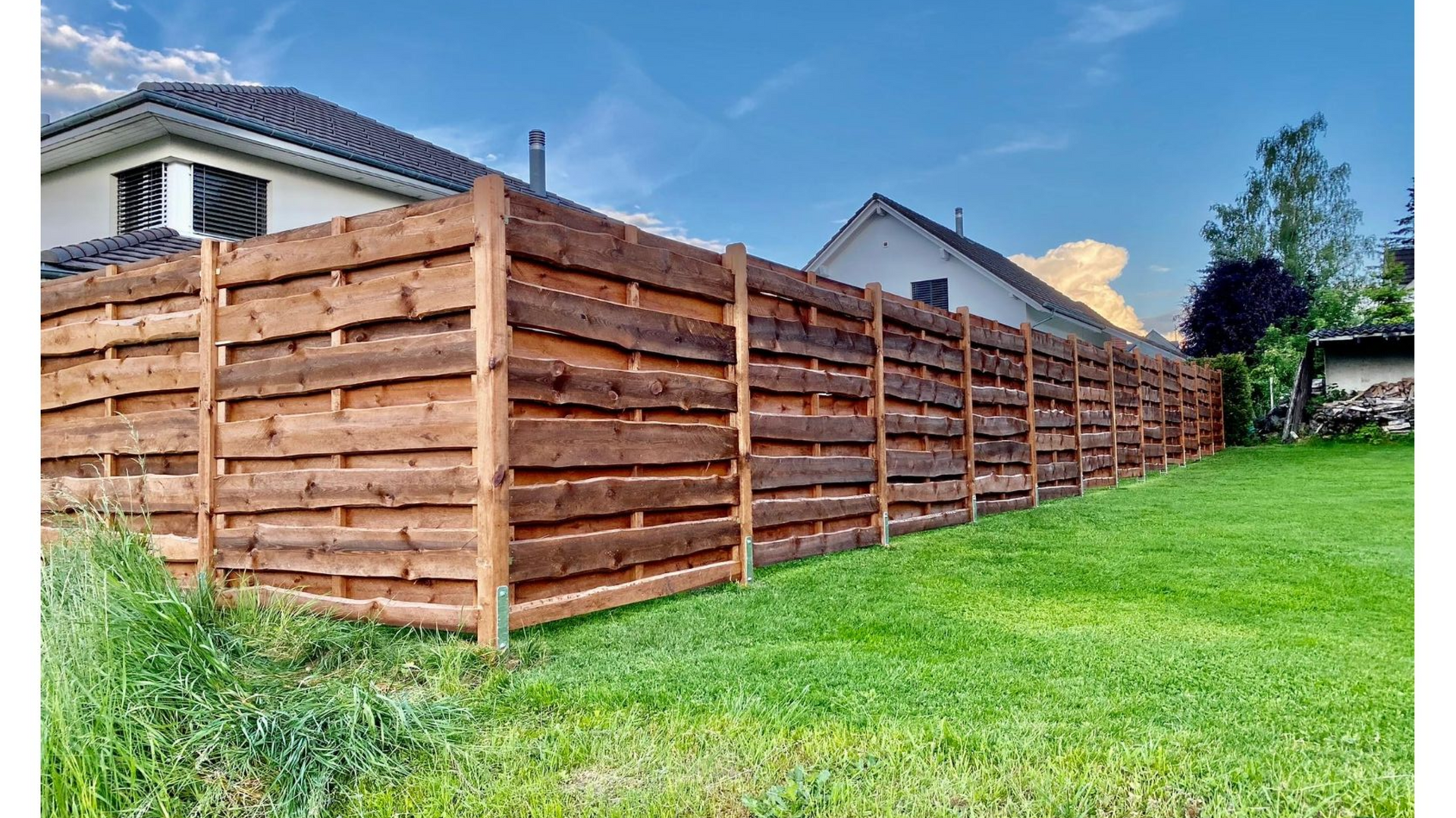 Sichtschutzzaun aus Holz in einem grasbewachsenen Hof, mit Häusern und einem blauen Himmel im Hintergrund.