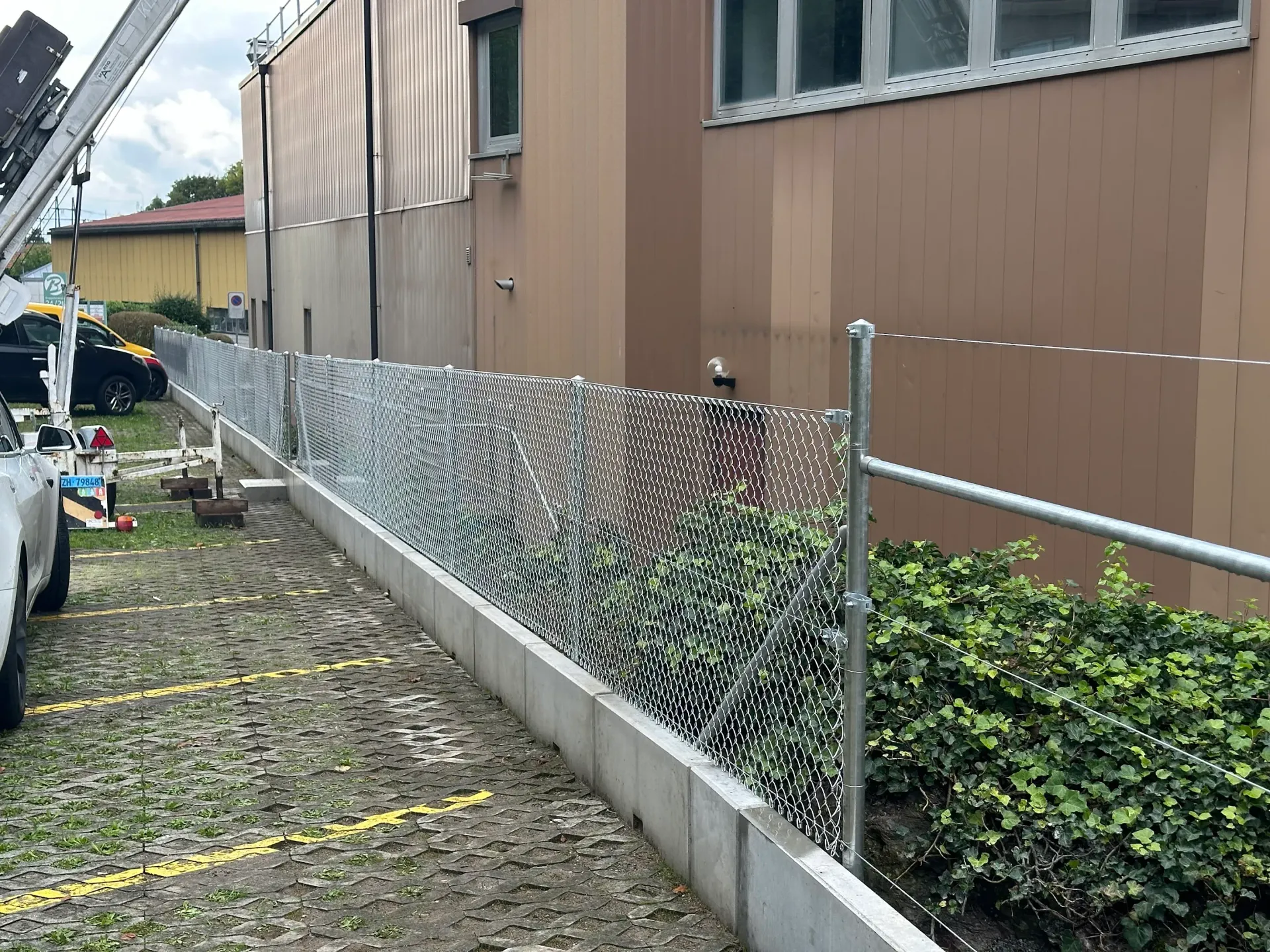 Maschendrahtzaun mit Sichtschutzlamellen entlang eines Gebäudes. Grau- und Grüntöne; Außenanlage.