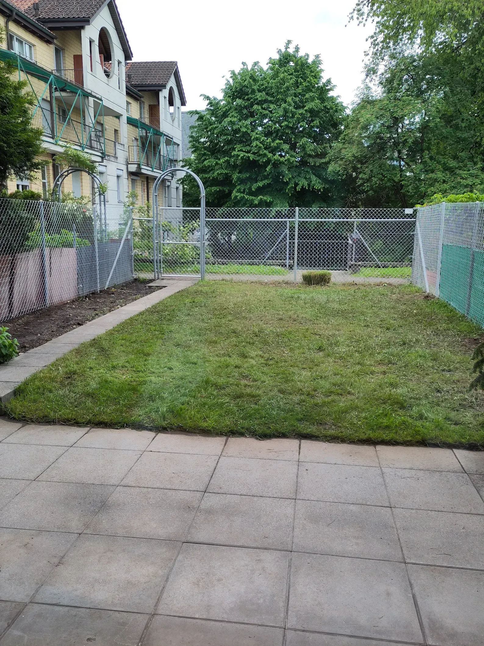 Ein grasbewachsener Hinterhof mit einer Terrasse, einem Zaun und einem Gebäude im Hintergrund.