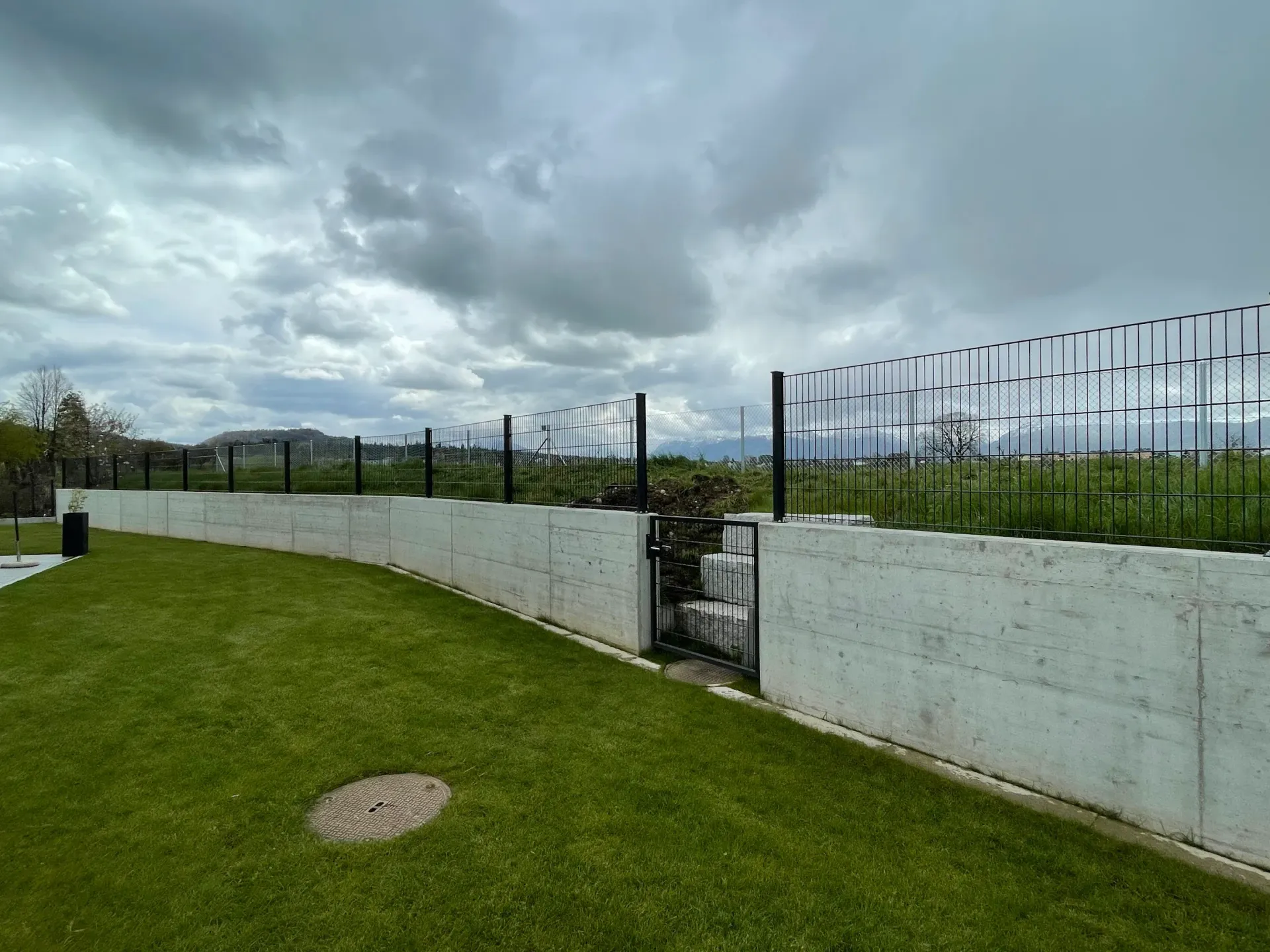 Grüner Rasen mit Betonmauer und dekorativem schwarzen Metallzaun unter bewölktem Himmel.