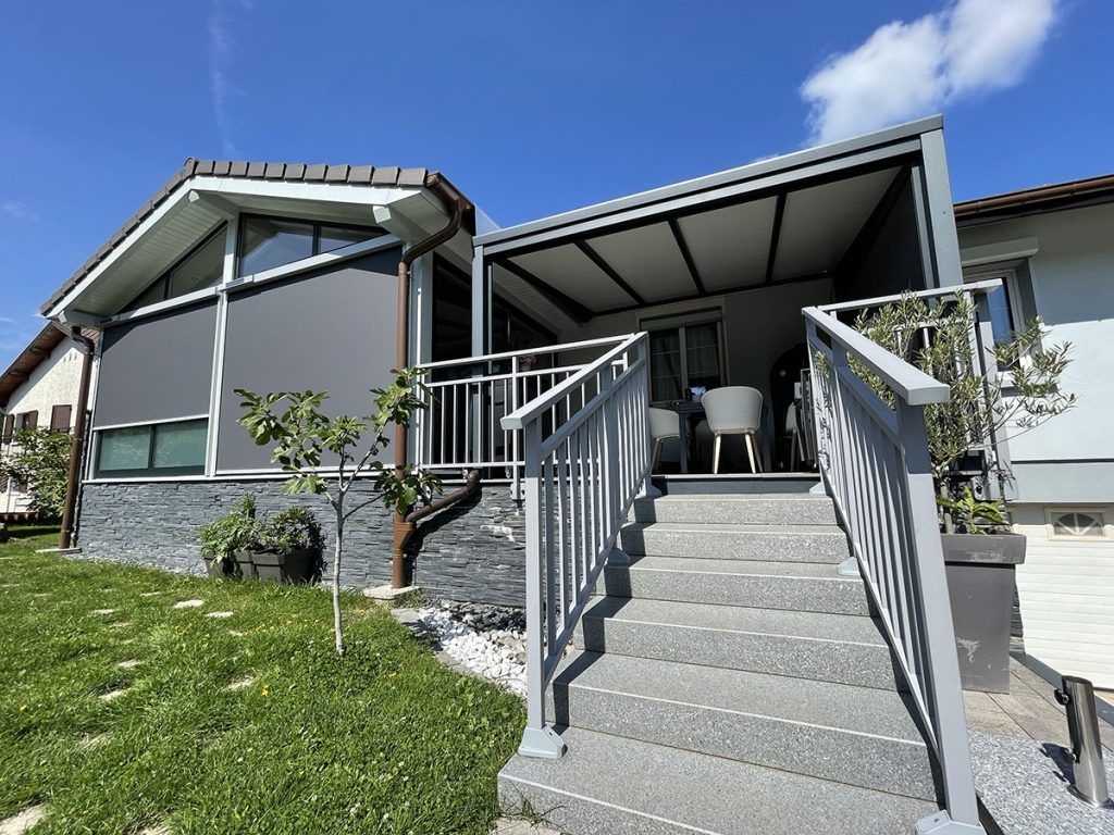 Extérieur d'une maison moderne avec marches d'entrée, balustrades en métal et porche couvert sous un ciel bleu.