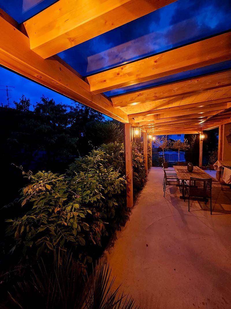 Patio couvert au crépuscule, éclairé par une douce lumière orangée, sol en pierre, table à manger et végétation luxuriante sous un ciel bleu.