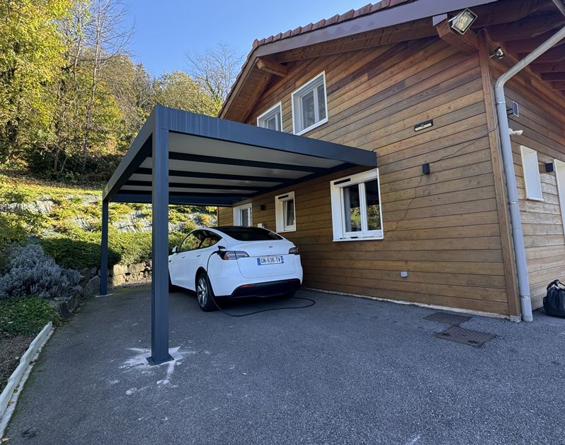 Voiture blanche garée sous un abri métallique moderne, à côté d'une maison en bois, sur une allée pavée.