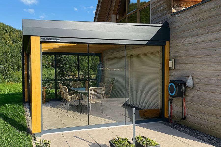 Terrasse vitrée attenante à une maison en bois, avec table et chaises sous un toit ombragé
