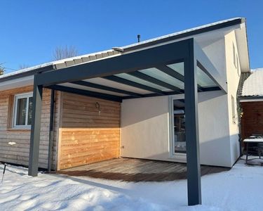 Abri de voiture enneigé avec des poutres de soutien sombres, attaché à une maison sous un ciel bleu